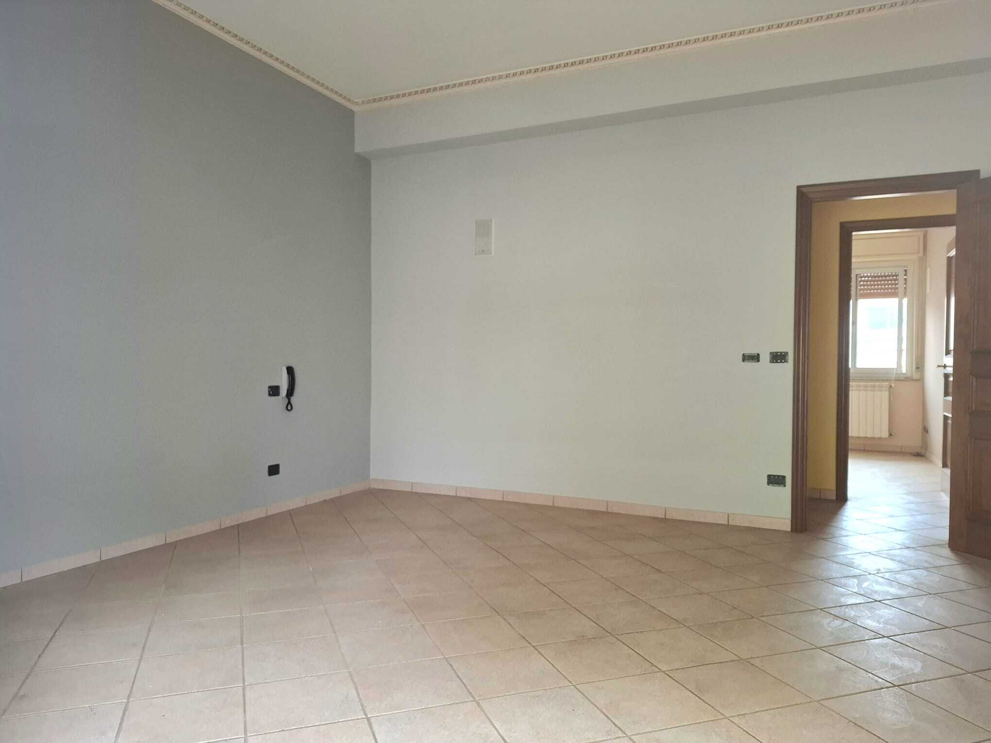 Appartamento vuoto con pareti bianche e pavimento in piastrelle beige, pronto per essere arredato, con accesso a una cucina luminosa e finestra che permette l'ingresso di luce naturale.