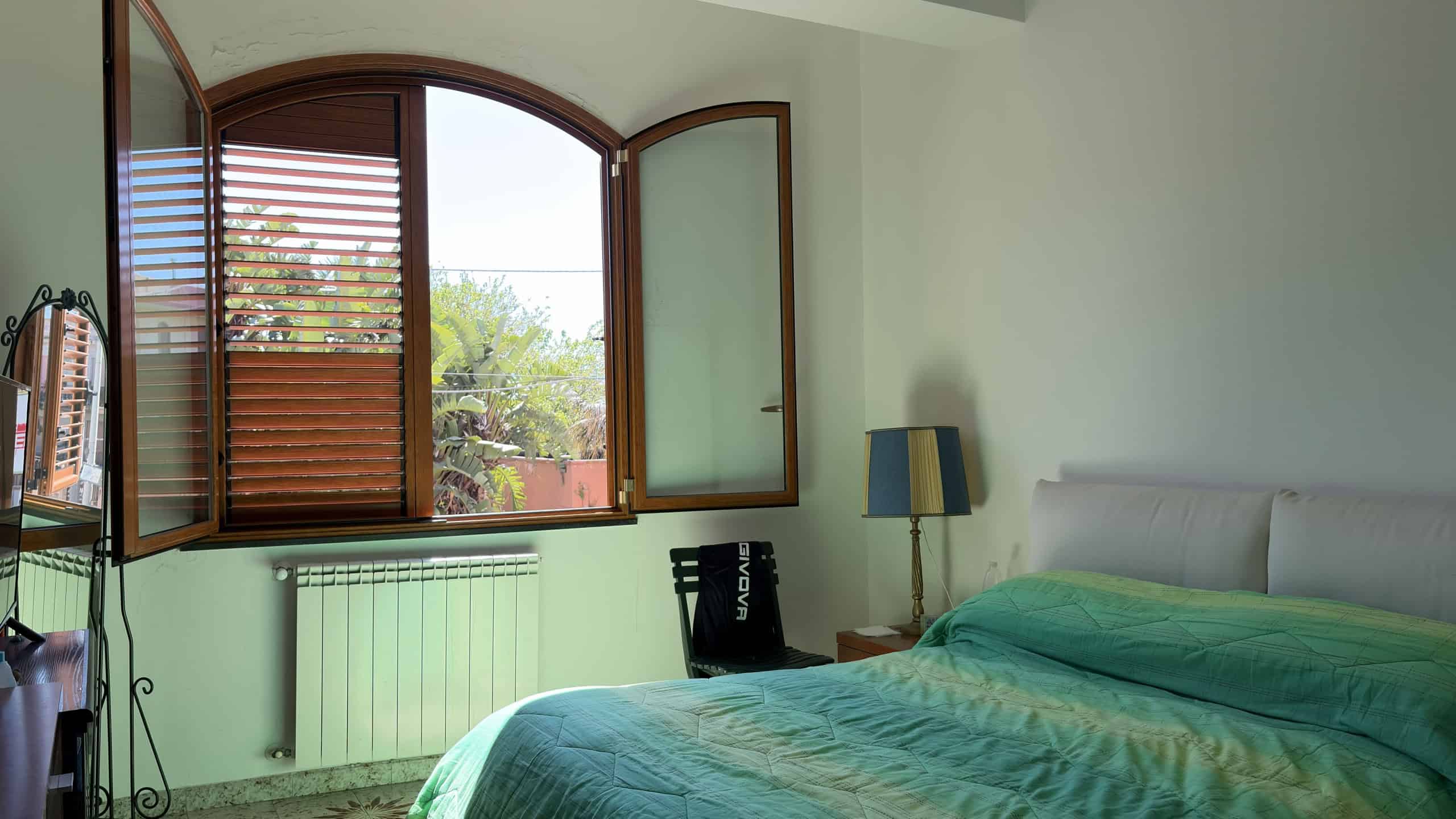 Ampia camera da letto con finestra ad arco, tende di legno e vista sul giardino, arredamento semplice e luminoso, ideale per ambienti residenziali di alta qualità.