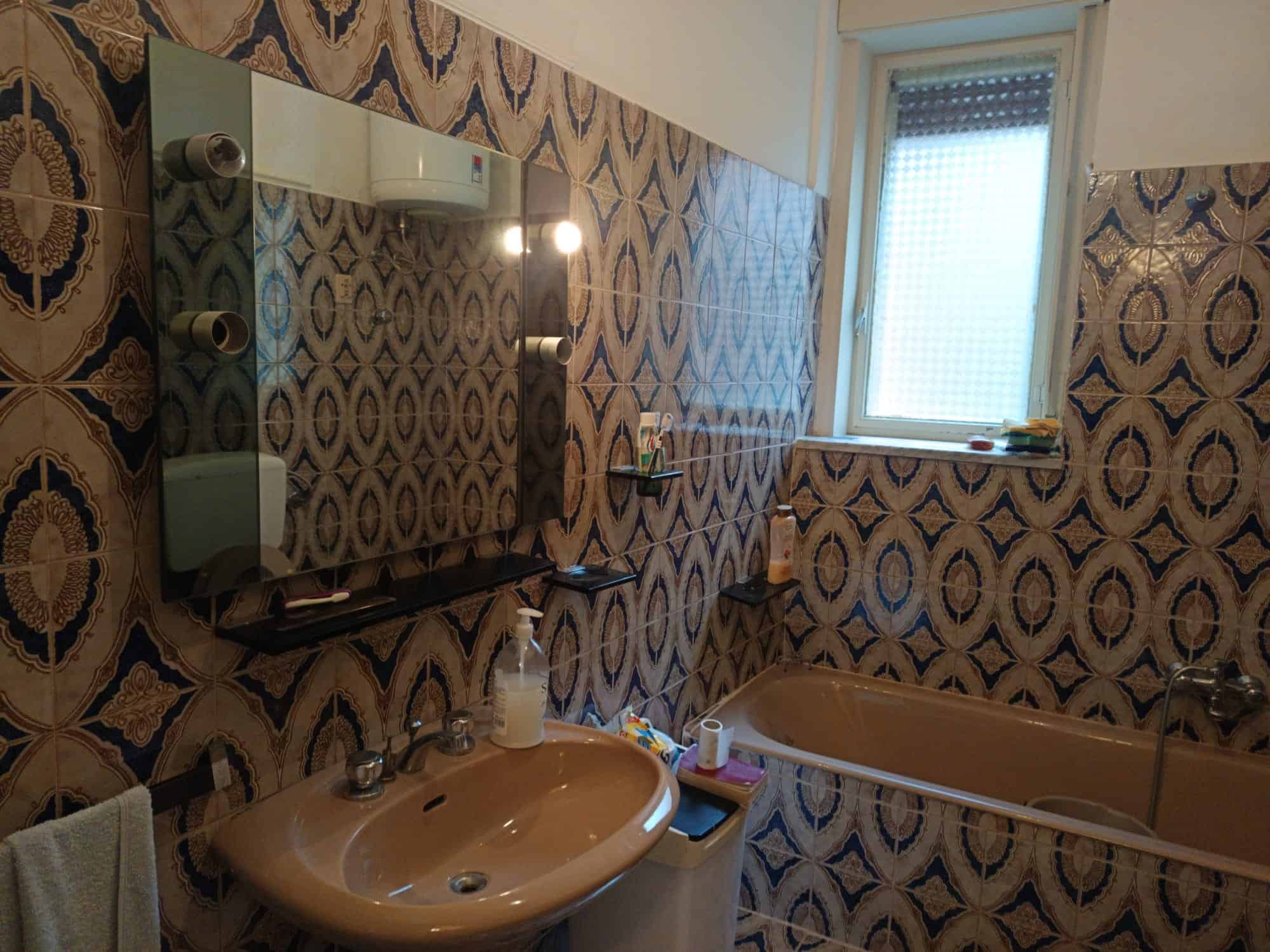 Specchio e lavabo in bagno con piastrelle decorative, finestra luminosa e vasca da bagno, ambiente accogliente e ben illuminato.