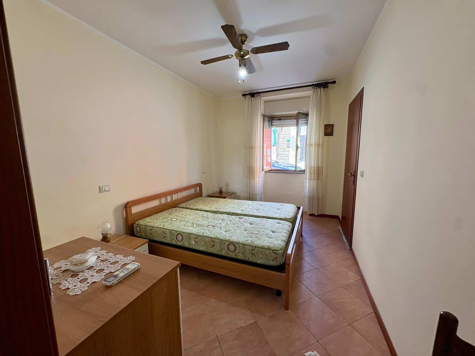 Letto matrimoniale con due materassi in una stanza luminosa e semplice, dotata di finestra, ventilatore a soffitto e arredi minimalisti. Ideale per soggiorni confortevoli e funzionali.