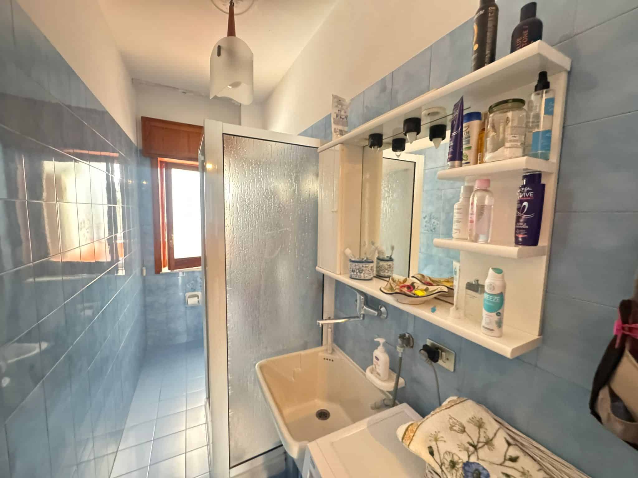Lavabo e specchio in bagno con pareti azzurre e scaffale con prodotti per la cura personale, illuminato da luce naturale proveniente dalla finestra.