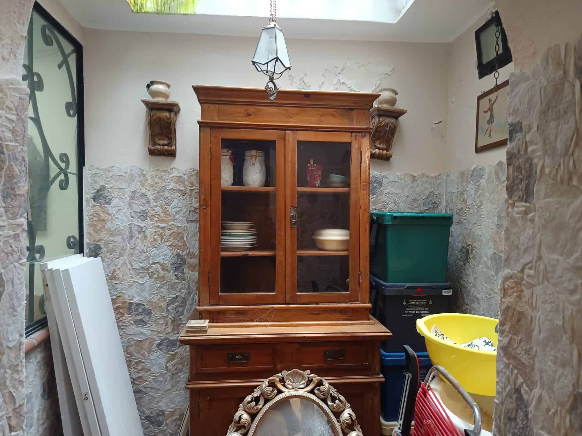 Riproduzione di un mobile antico in legno con vetrina, collocato in un ambiente domestico con pareti in pietra e decorazioni artistiche. Perfetto per arredamento classico e rustico, ideale per conserv.