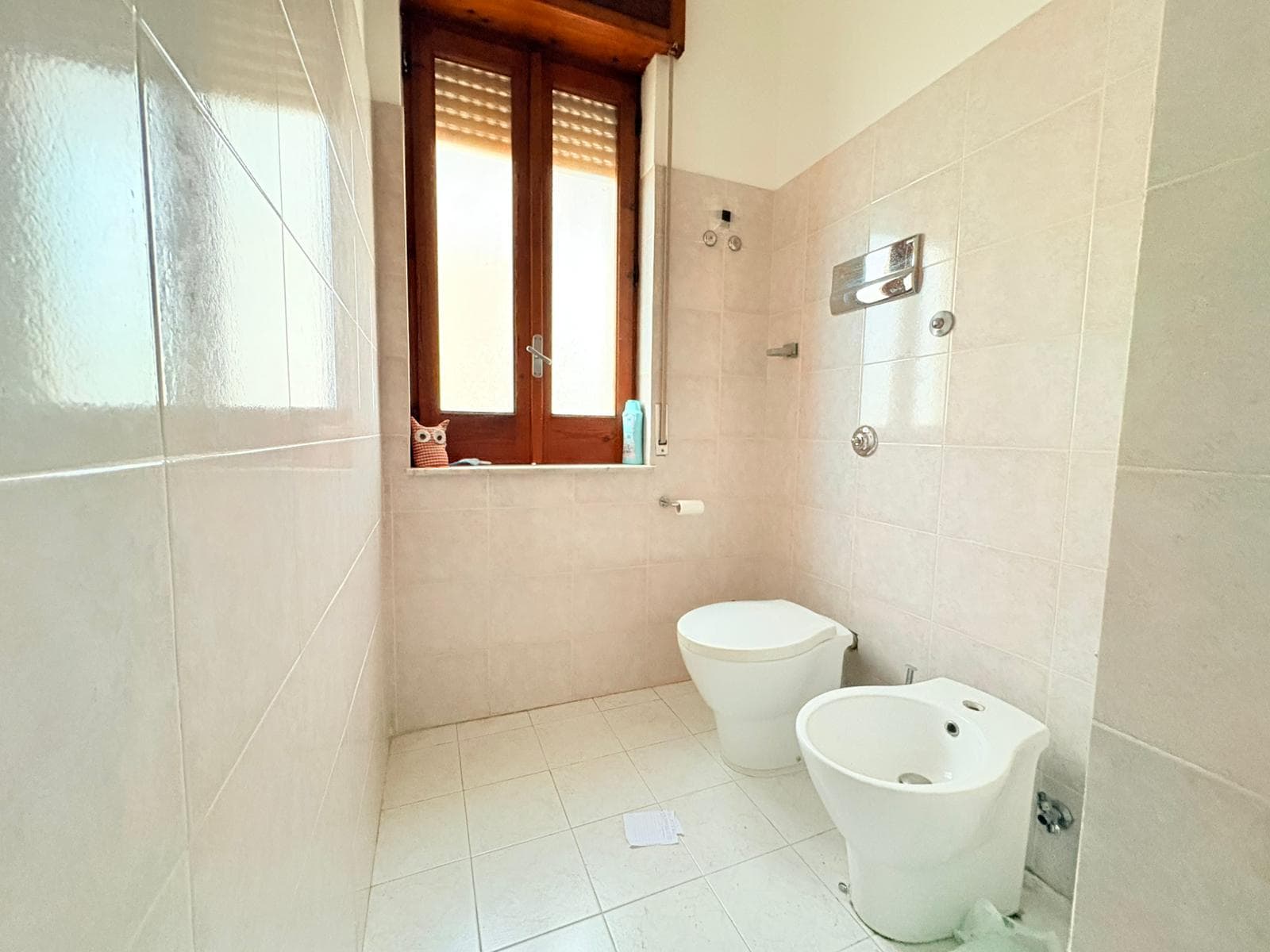 Pulsante di bagno moderno con WC e bidet, finestra in legno che permette l'ingresso di luce naturale, pareti e pavimenti in piastrelle chiare, ideale per ambienti di servizi igienici funzionali e lumi.