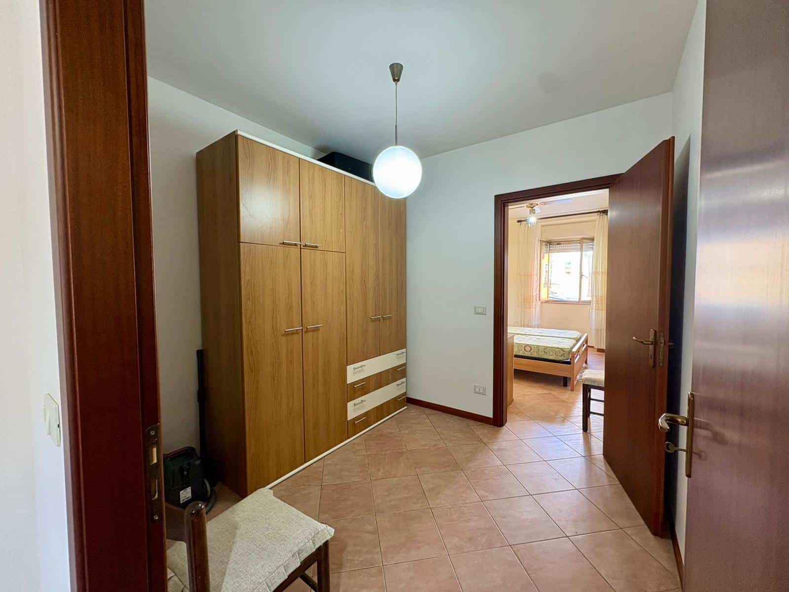 Aggiungi un armadio in legno in un ingresso di casa con porta aperta che conduce a una camera da letto, illuminata da una lampada a soffitto e con pavimento in piastrelle di ceramica.