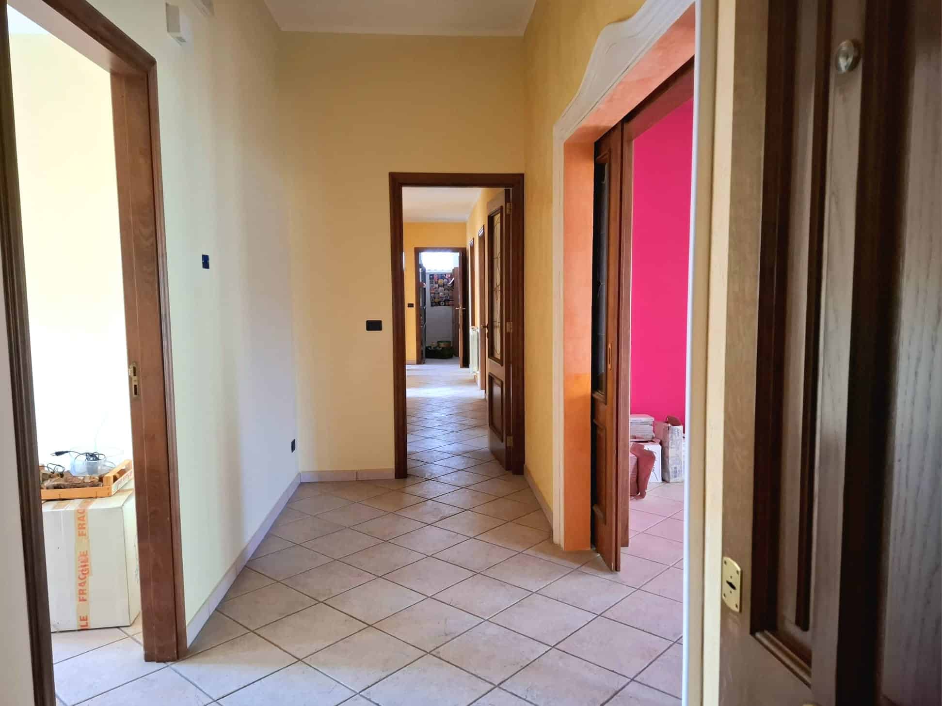 Ampia vista di un corridoio interno di una casa con pareti color crema e porte in legno, illuminato dalla luce naturale, che conduce a diverse stanze e spazi abitativi.