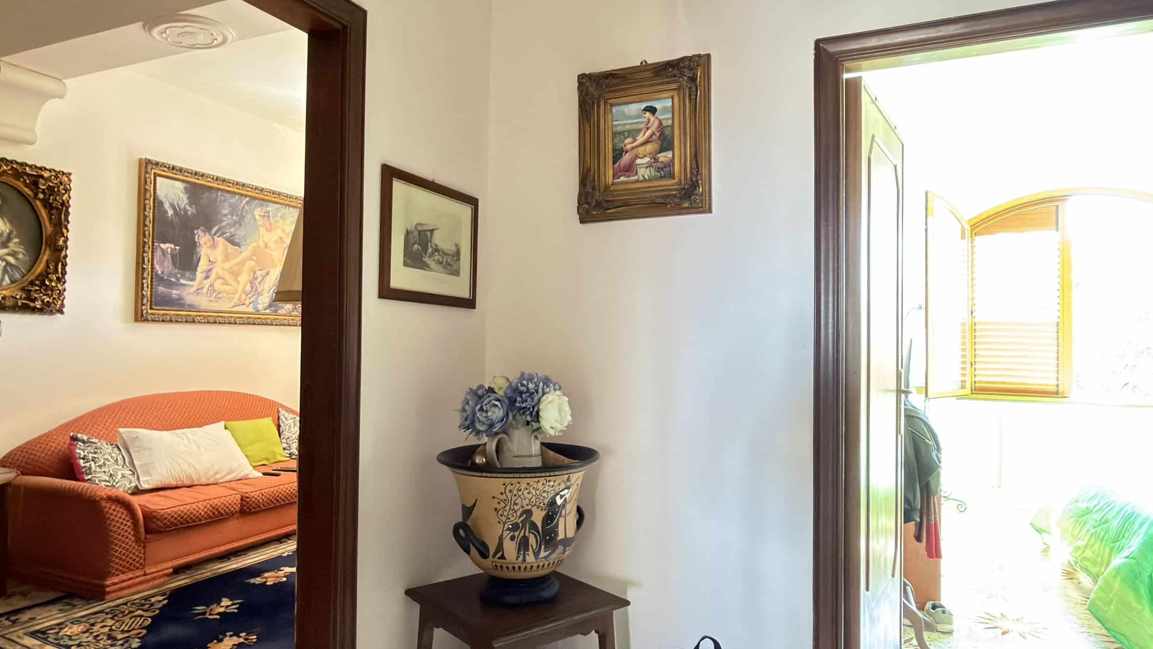 Elegante ingresso con decorazioni classiche, vaso di ceramica con fiori e dipinti alle pareti, illuminato da luce naturale proveniente da una porta aperta che conduce a un'area luminosa e accogliente.
