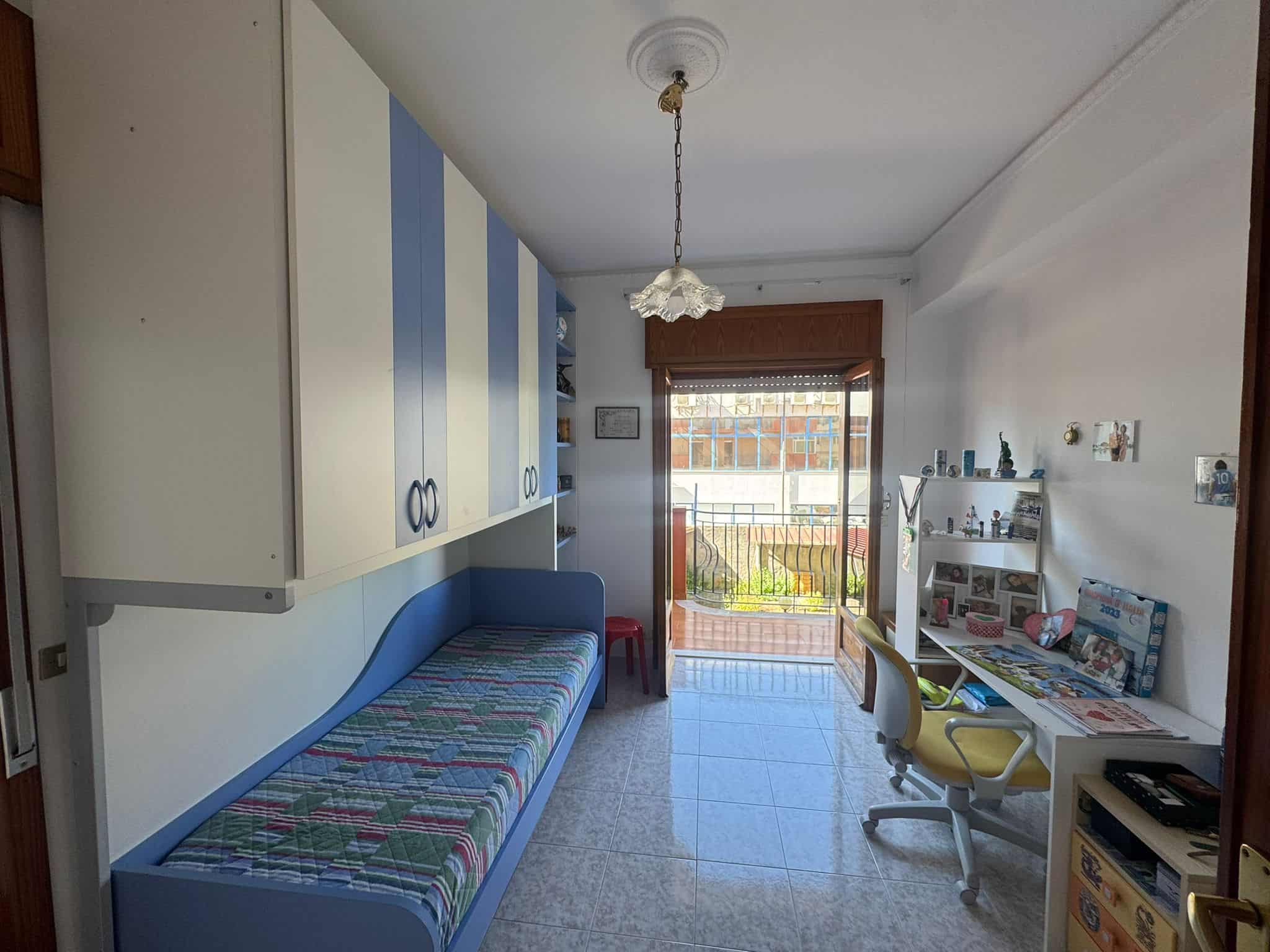 Sedile a letto in una stanza luminosa con armadiature bianche e una scrivania con sedia gialla, vista su un balcone con ringhiera e piante, ambiente accogliente e ben organizzato.
