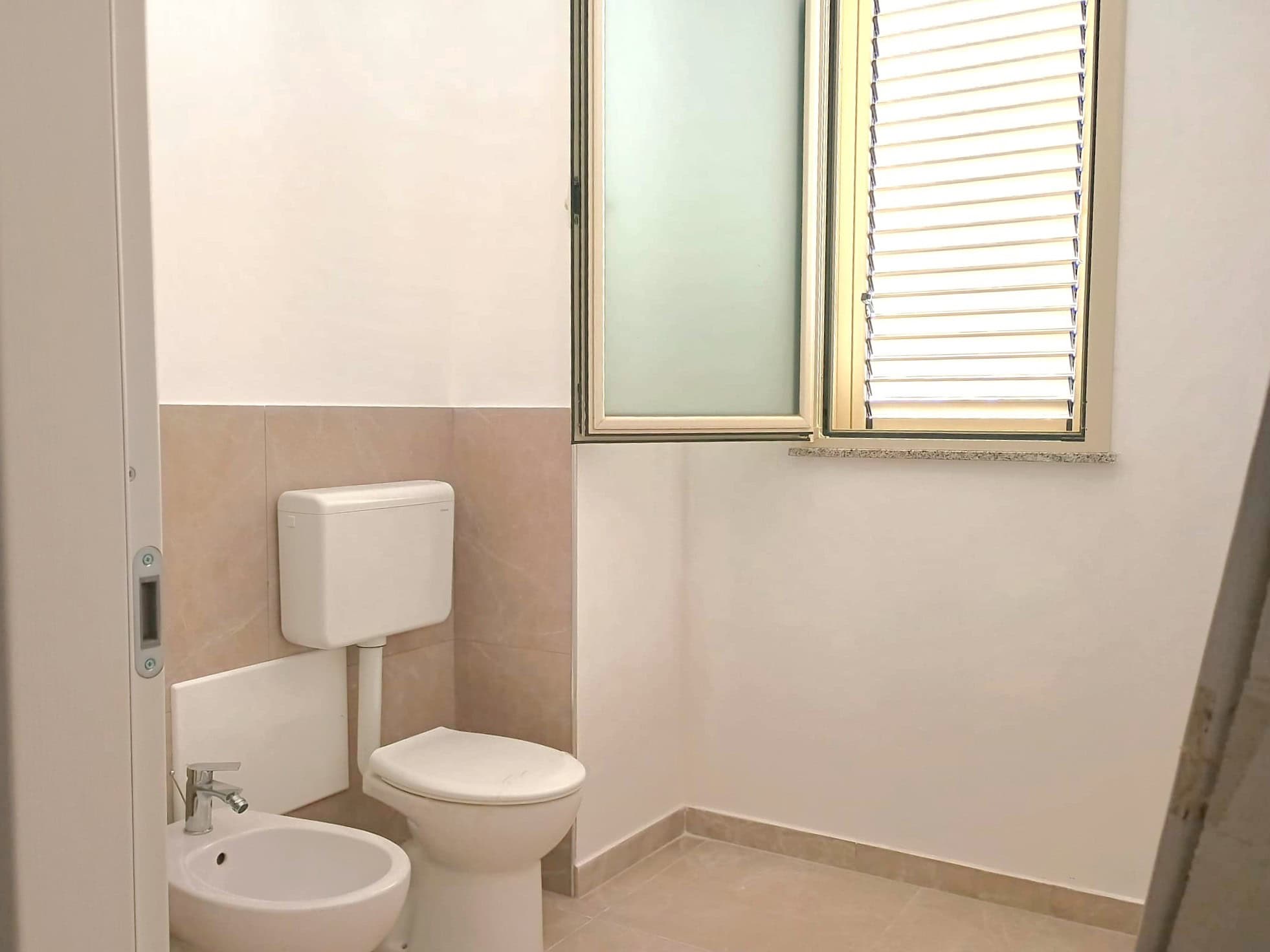 Serratura di sicurezza per bagno con WC e bidet in un ambiente luminoso e pulito, ideale per appartamenti o strutture ricettive. Finestra con persiane per ventilazione e luce naturale.