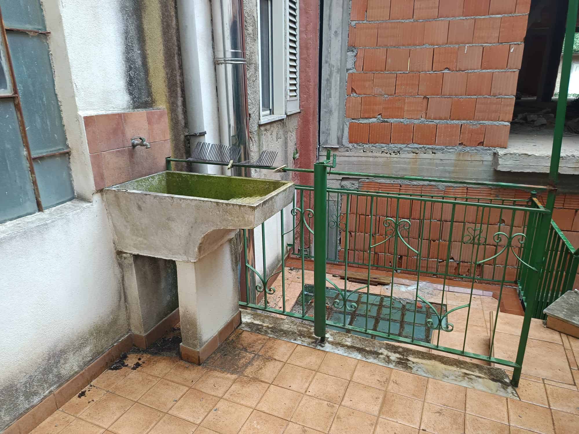 Lavandino esterno in pietra con area di lavaggio, vicino a pareti in mattoni e cemento, con ringhiera verde che delimita un'area di accesso. Ideale per uso domestico e attività di giardinaggio.
