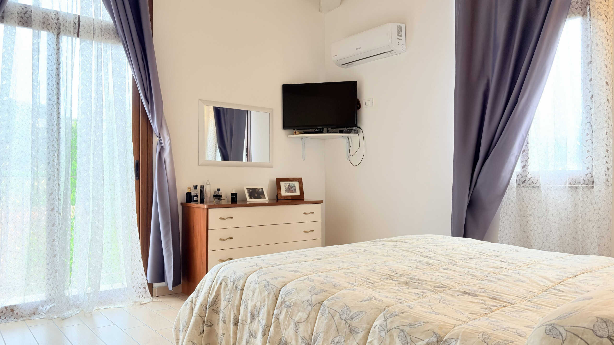 Ampio soggiorno con pareti bianche, aria condizionata, TV a schermo piatto, specchio e mobile con decorazioni, illuminato da grandi finestre con tende viola e tende leggere, ideale per relax e comfort.