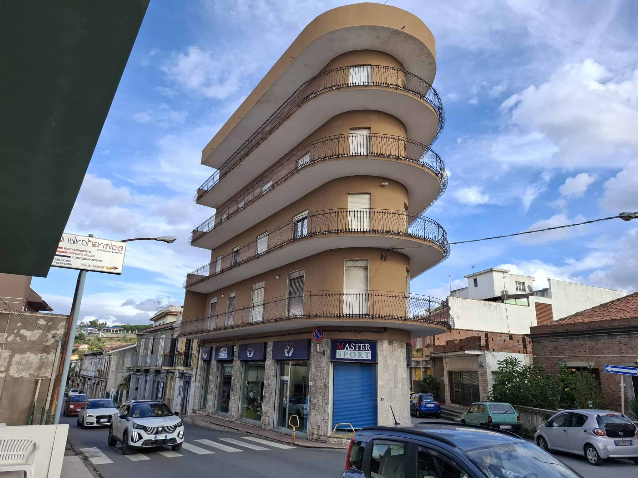 Il palazzo moderno con balconi curvi e facciata in tonalità beige, situato in un'area urbana con negozi al piano terra e veicoli parcheggiati lungo la strada, sotto un cielo sereno con nuvole sparse.