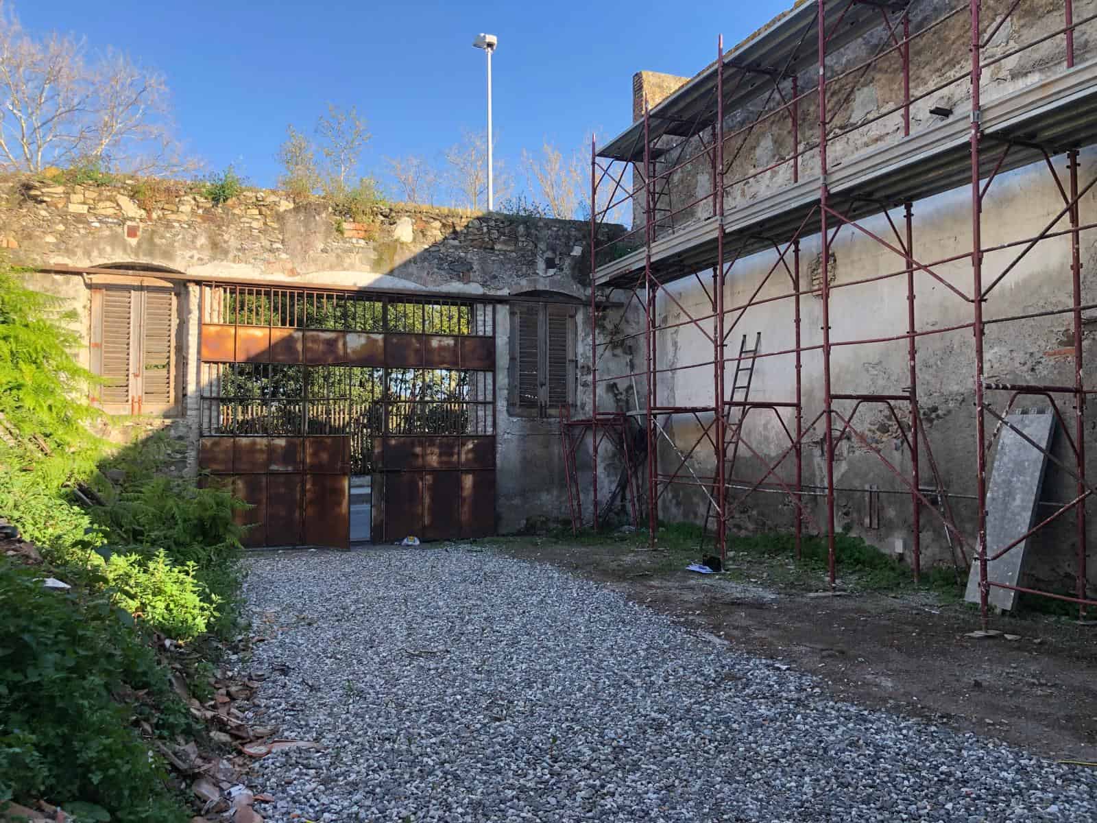Il cancello in ferro battuto e il muro di pietra di un edificio storico in fase di ristrutturazione, con impalcature e lavori di restauro, sotto un cielo azzurro.