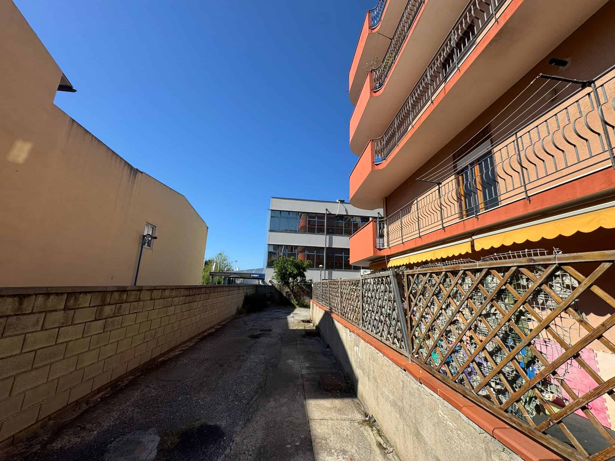 Viale residenziale con edifici moderni e balconi, strada stretta e cielo sereno, vista da un'angolazione laterale.