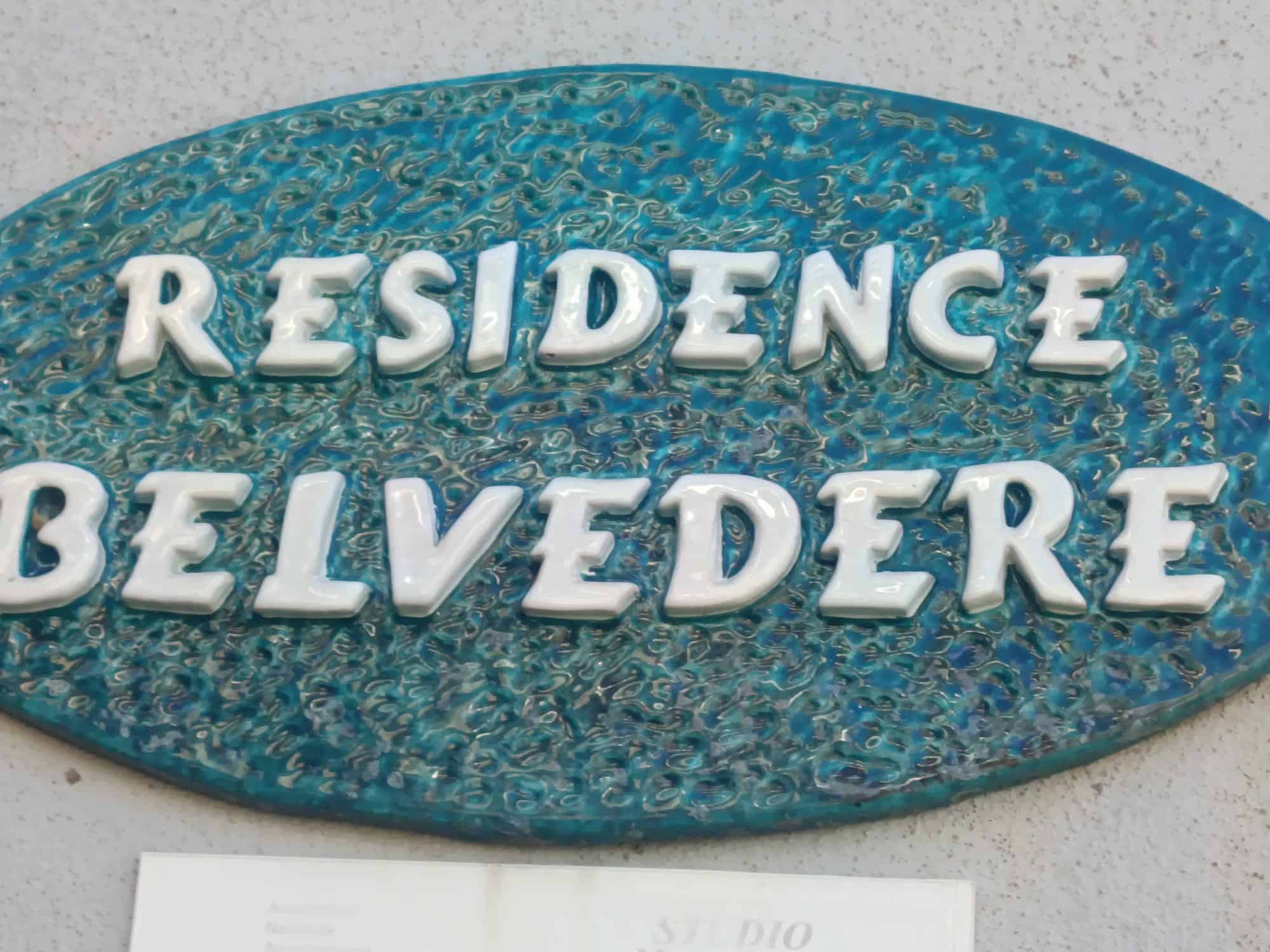 Pannello in ceramica con scritta "Residence Belvedere" su parete esterna, decorato con dettagli blu e bianco.