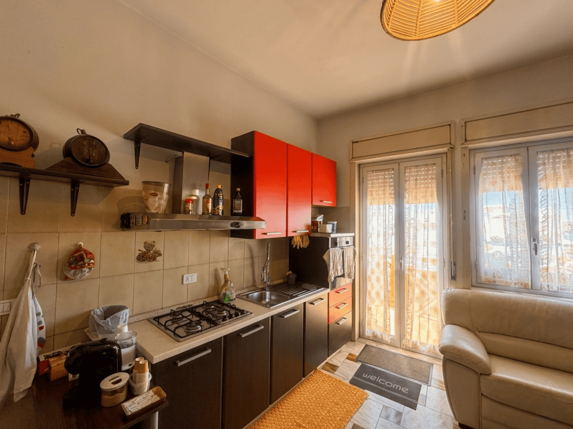 Ampio angolo cottura con mobili moderni, pareti beige e accessori pratici, illuminato dalla luce naturale proveniente dalle finestre e dalla porta finestra con tende trasparenti, ideale per vivere in.