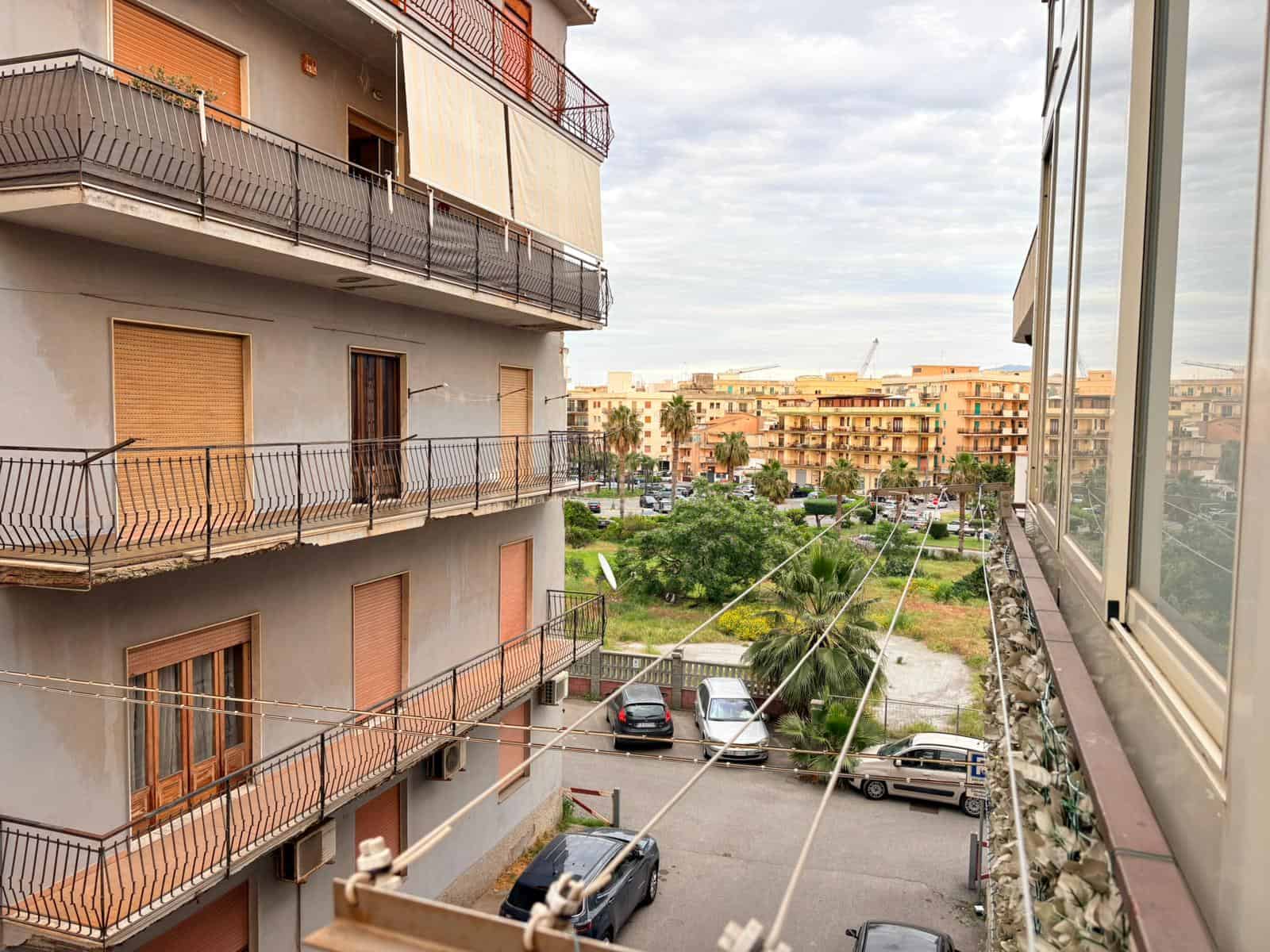 Balcone di un appartamento con vista su un giardino condominiale e edifici residenziali in lontananza, in un contesto urbano italiano.
