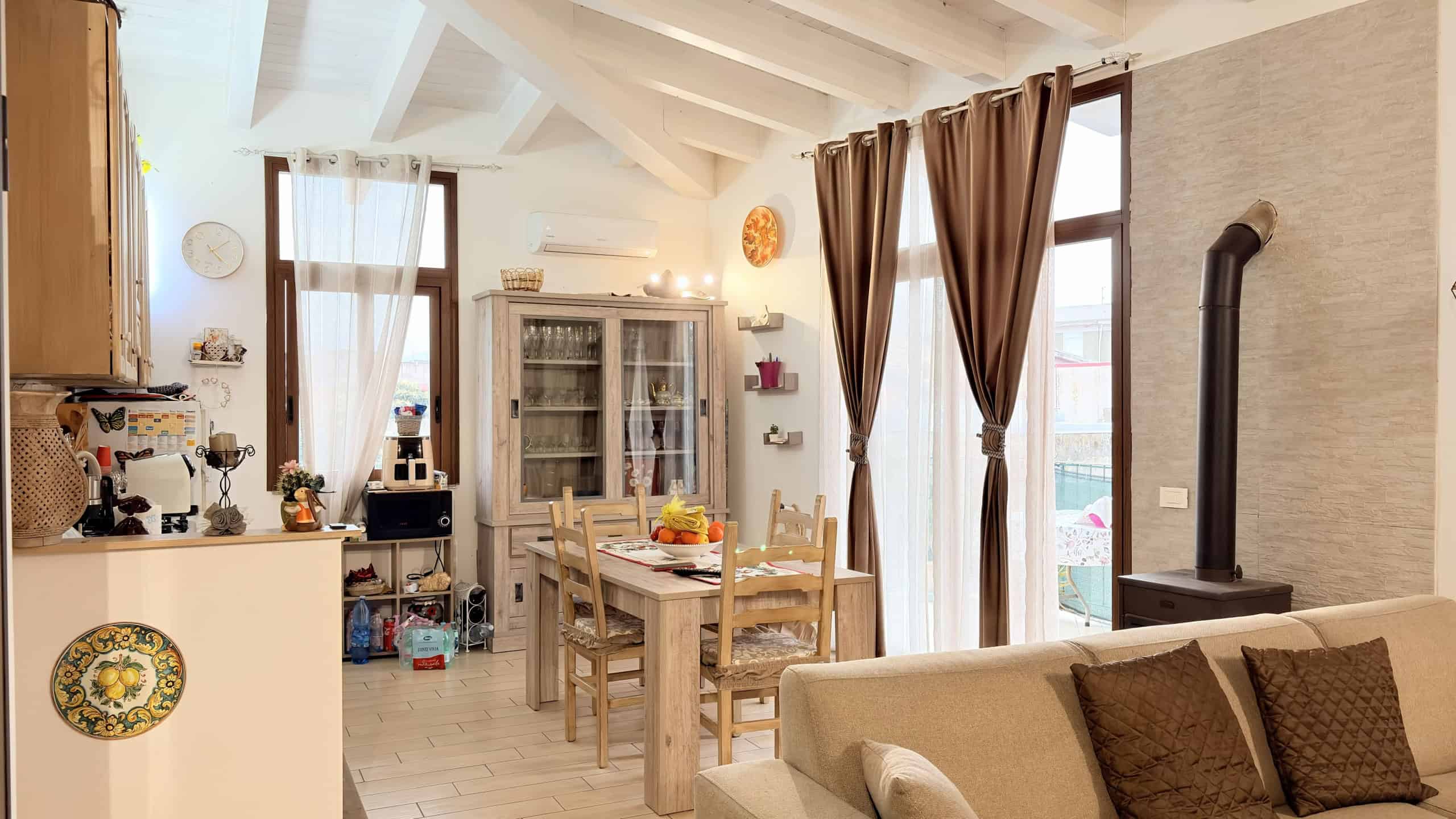 Ampia zona living con cucina aperta, tavolo da pranzo in legno, divano beige e camino moderno, illuminata da grandi finestre con tende marroni, in un appartamento luminoso e accogliente.