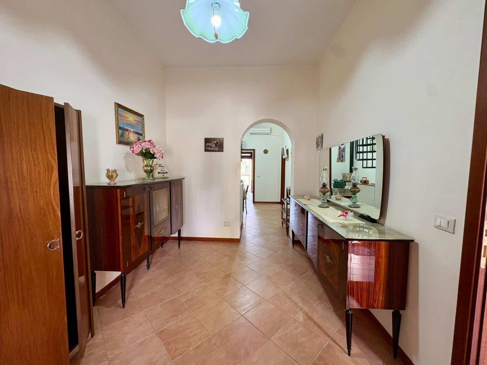 Corridoio spazioso con mobili in legno, decorazioni floreali e specchi, illuminato da lampada a soffitto, in un appartamento elegante e accogliente.