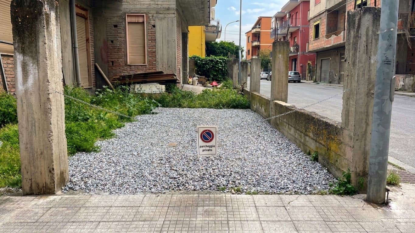 Auto in sosta non autorizzata in un'area di parcheggio all'aperto, delimitata da pali di legno e recinzione in cemento, in un quartiere residenziale con edifici colorati e strada asfaltata.