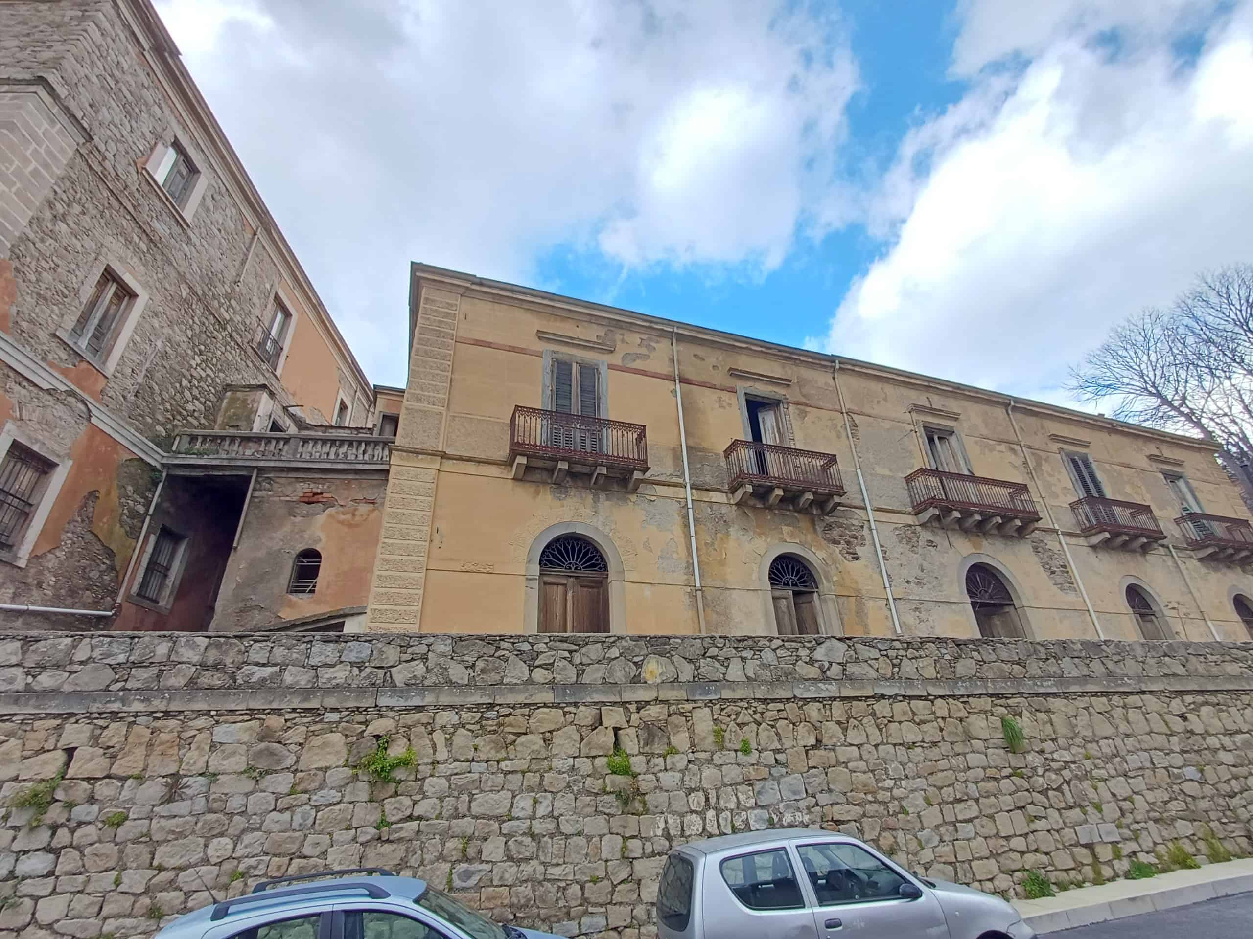 Edificio storico in pietra con balconi in ferro battuto e vista sul cielo azzurro, situato in un quartiere antico di città italiane, con auto parcheggiate davanti.