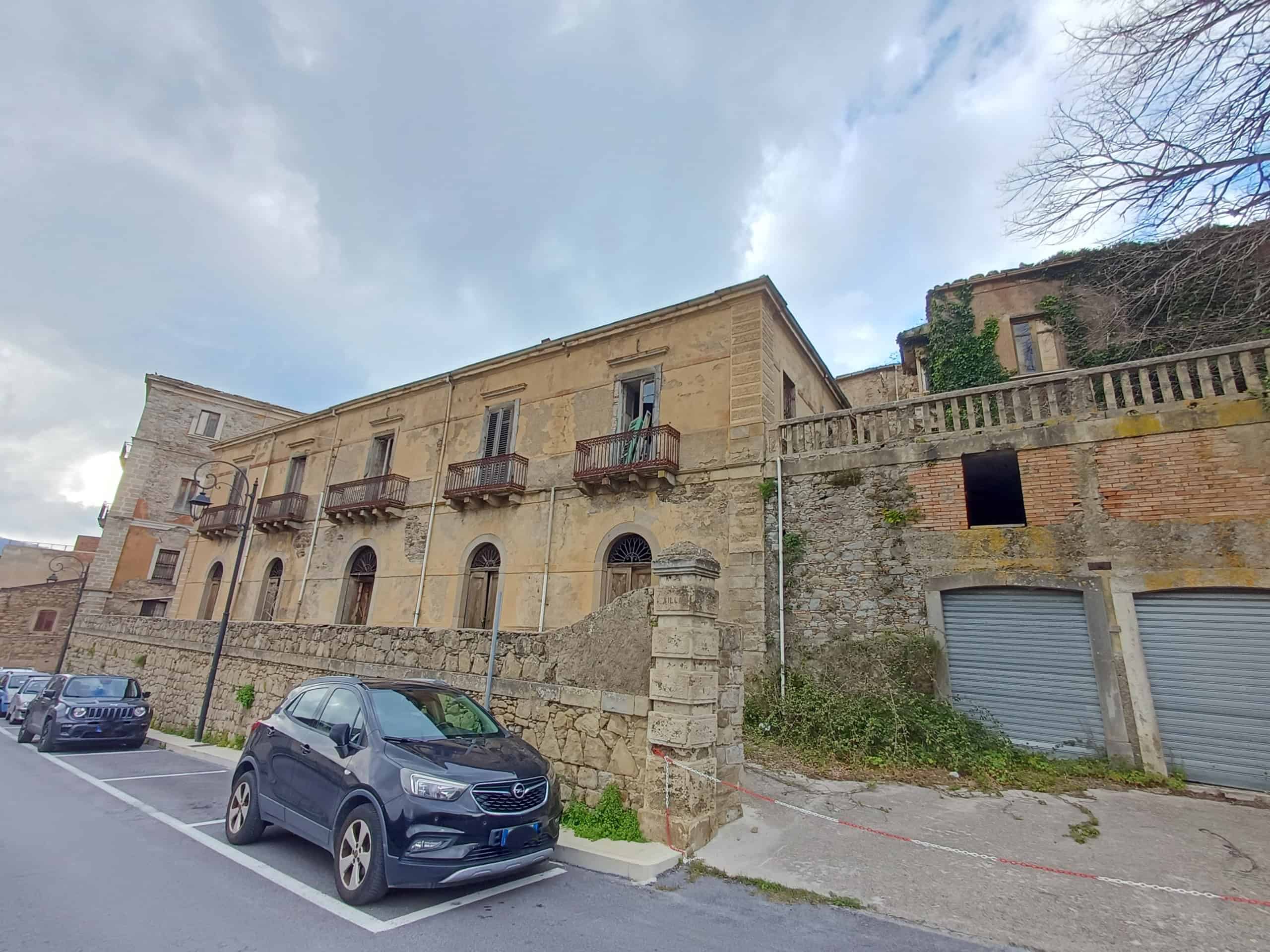 L'immagine mostra un edificio storico in stile classico con balconi in ferro battuto, situato in un'area urbana con parcheggio e auto moderne in primo piano, sotto un cielo nuvoloso.