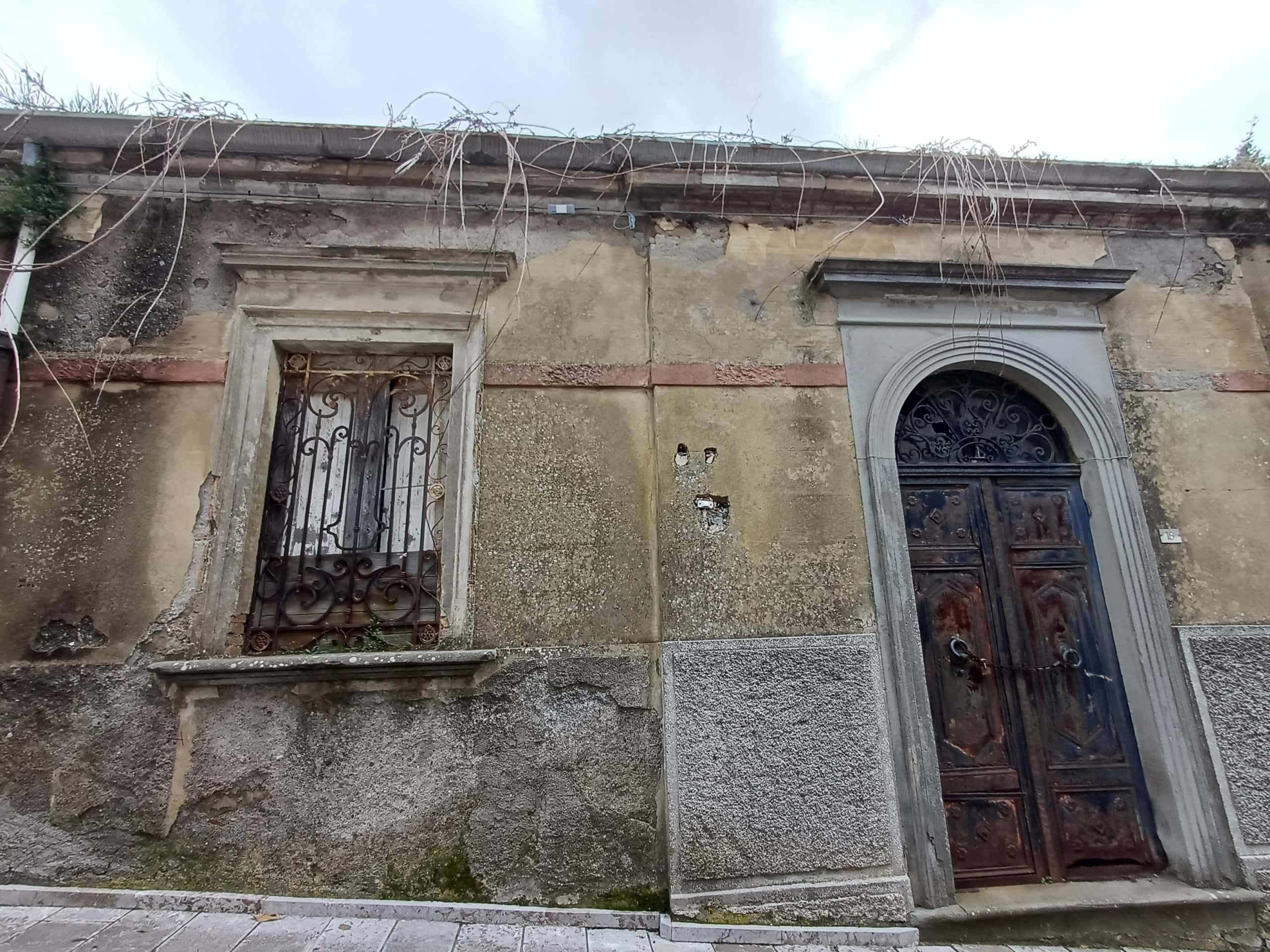 Vecchio edificio con finestra e porta in stile antico, pareti in pietra e dettagli decorativi in metallo, mostra segni di deterioramento e abbandono, ideale per interventi di restauro e valorizzazione.