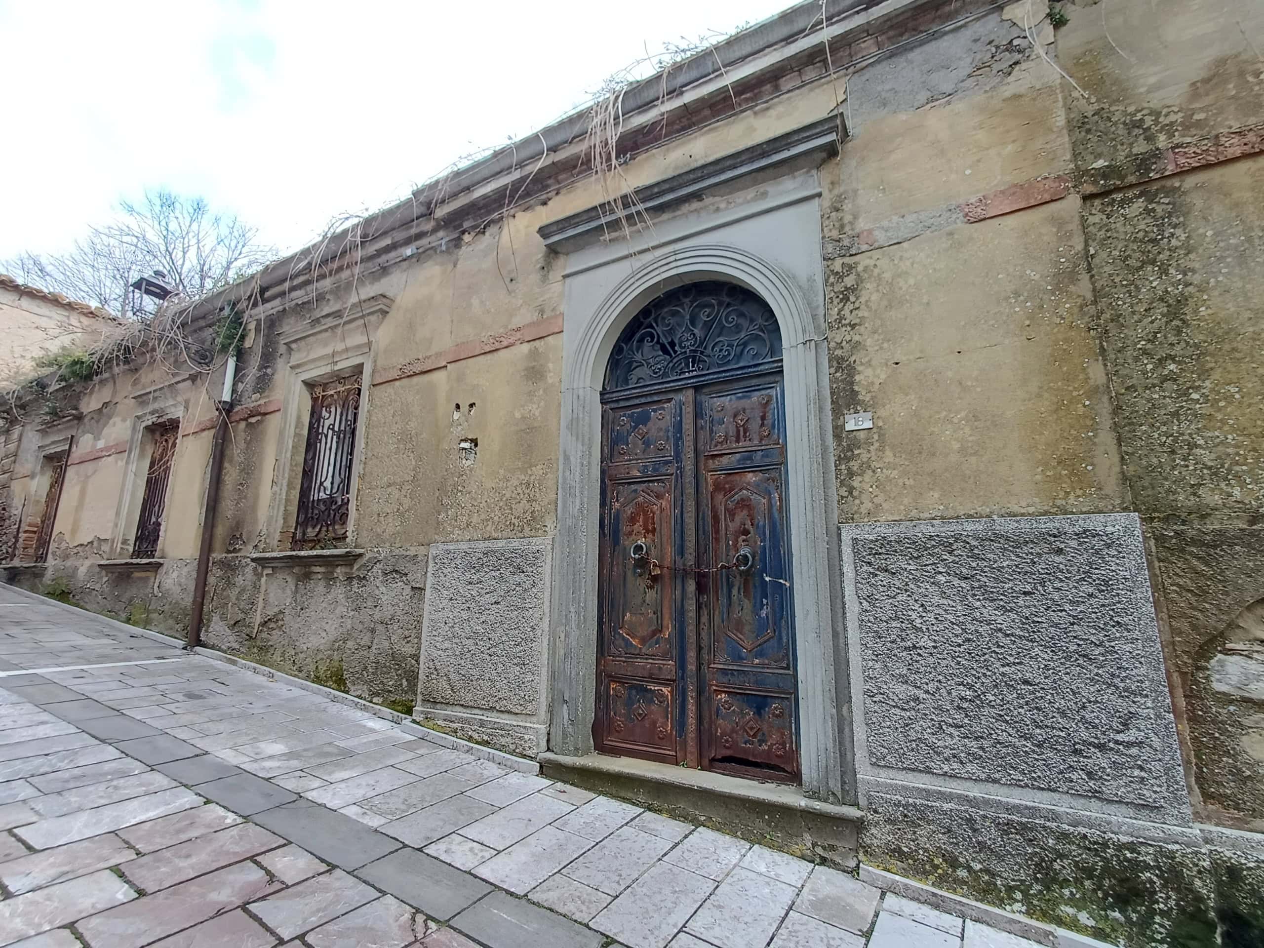 Porta d'ingresso in legno antico con dettagli decorativi e pareti in pietra, situata in una strada storica italiana.