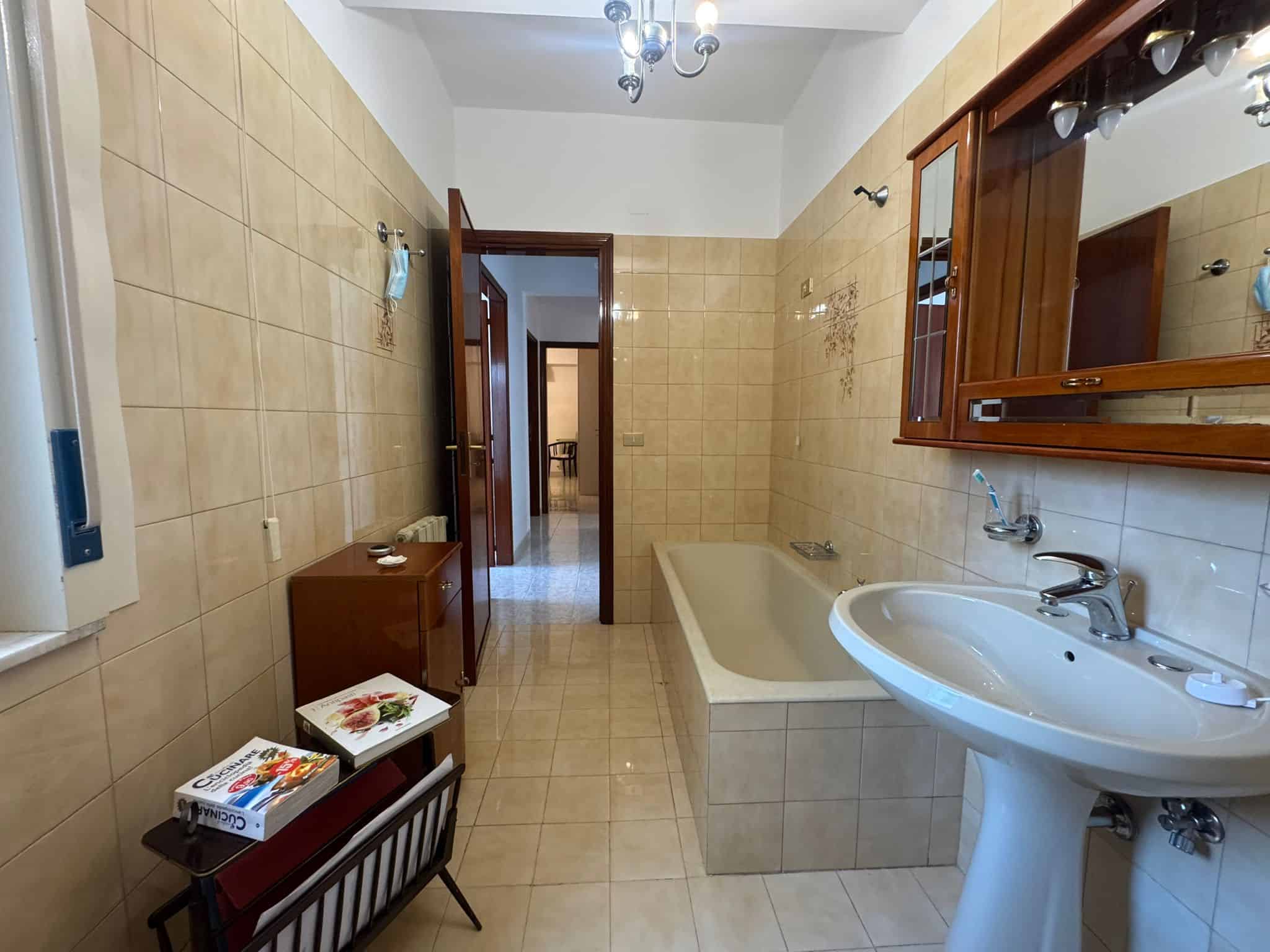 Ampia stanza da bagno con vasca, lavabo e specchio in legno, pareti rivestite di piastrelle beige, illuminata da luce naturale e lampadario centrale, vista verso la porta d'ingresso.