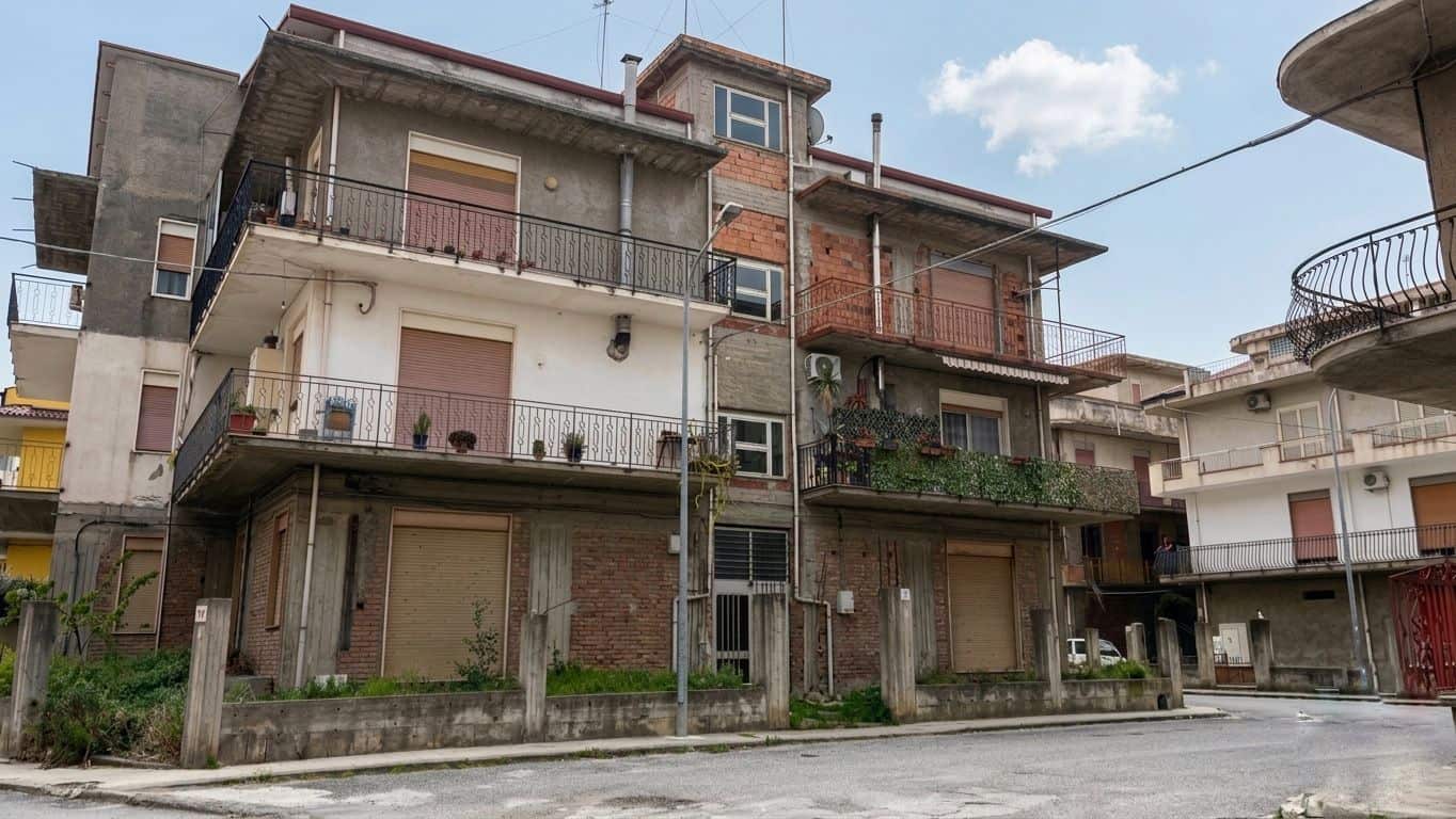 Balcone e facciata di un edificio residenziale in un quartiere urbano, con pareti in cemento e mattoni, balconi con ringhiere in ferro e piante sul balcone superiore, sotto un cielo sereno.