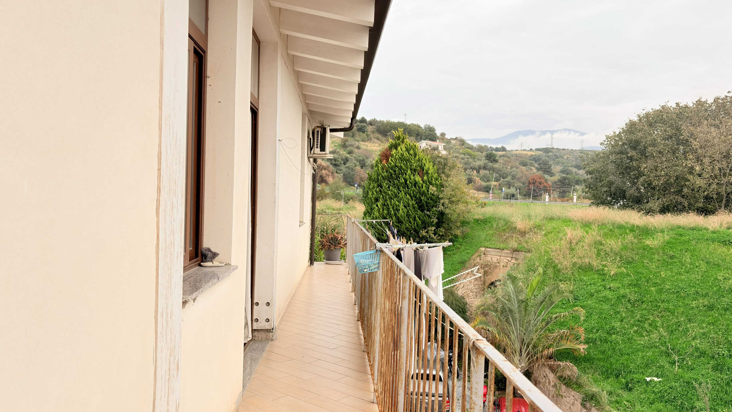Balcone di una casa con vista su un paesaggio verde e collinare, con piante e alberi, in una giornata nuvolosa, ideale per immagini di immobili e ambienti esterni.