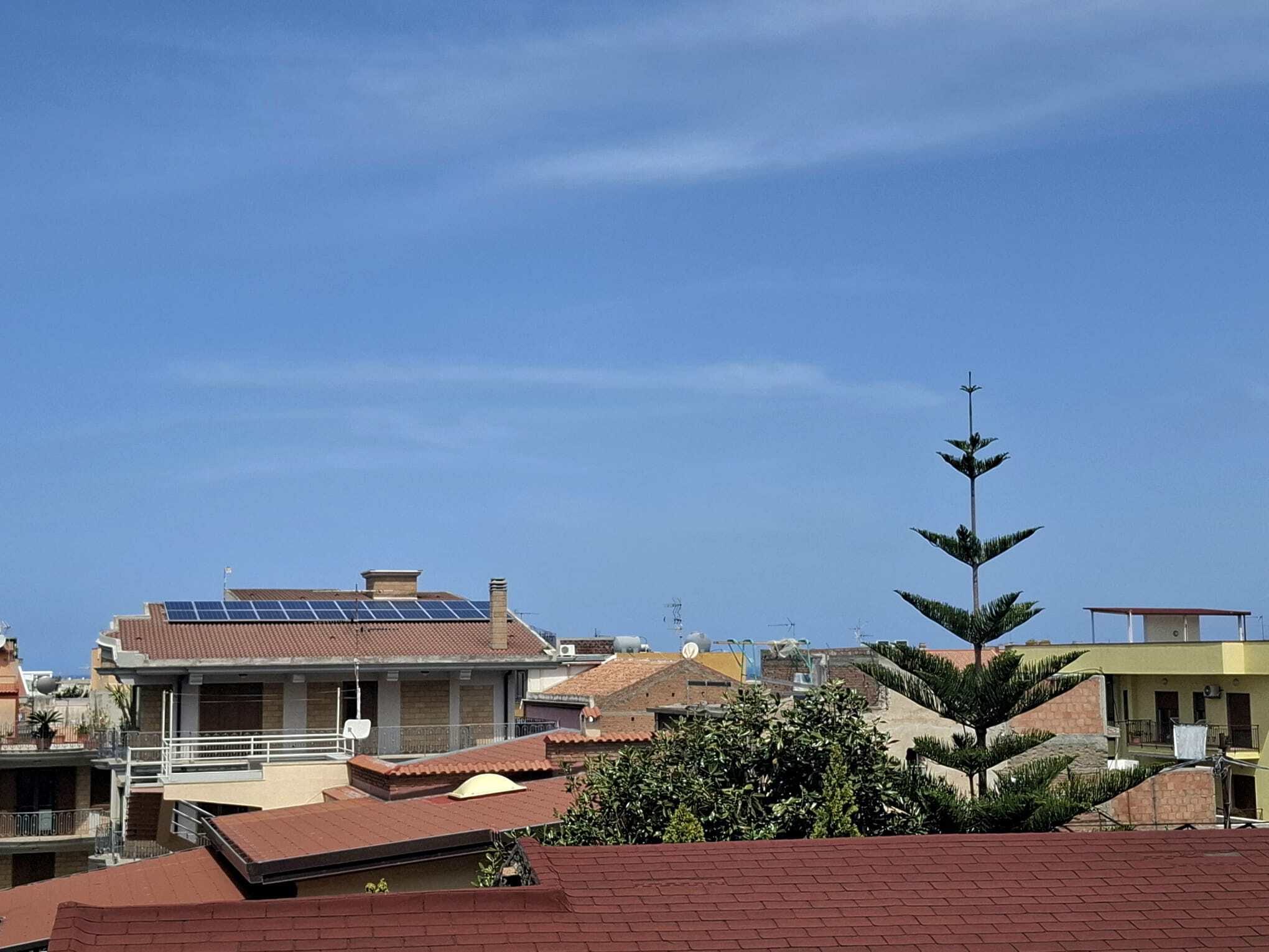 Pannello solare installato sul tetto di una casa residenziale in un quartiere urbano, con altre abitazioni e un albero di pino in primo piano, cielo sereno sullo sfondo.