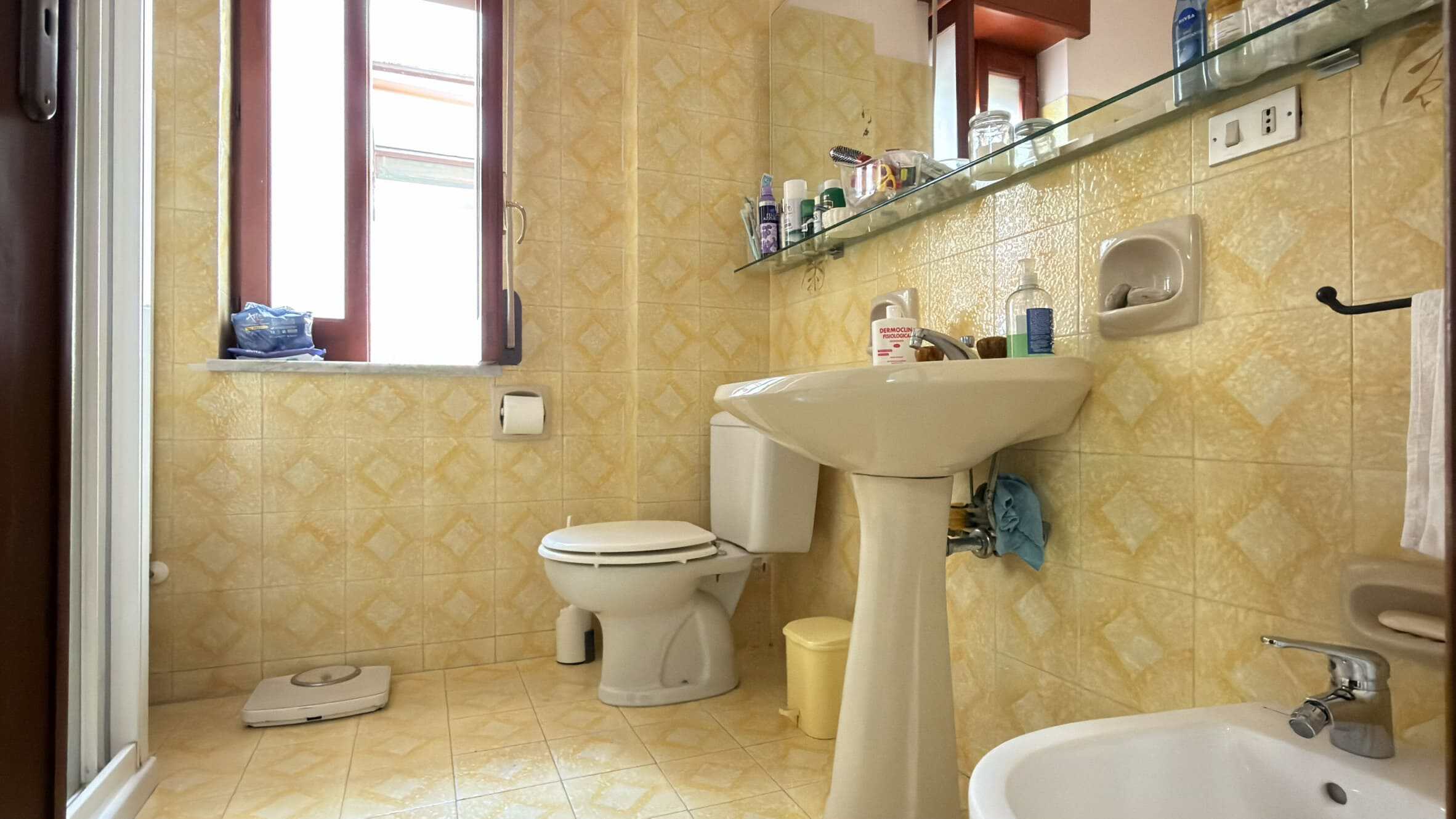 Ampia immagine di un bagno con pareti e pavimento in piastrelle gialle, lavabo, WC e bidet, con finestra che permette l'ingresso di luce naturale, accessori e prodotti per l'igiene.