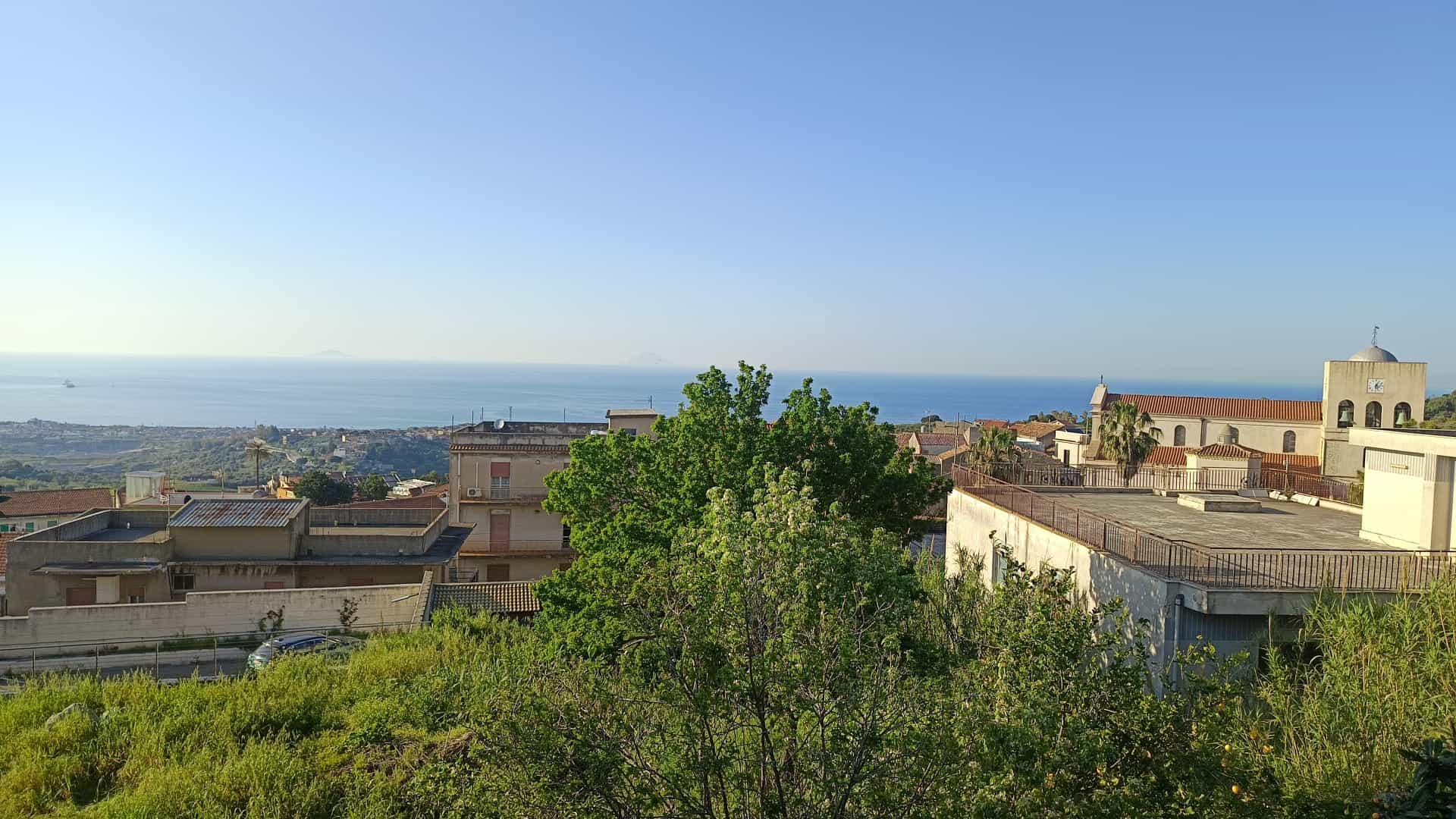 Vista panoramica sulla città con il mare sullo sfondo, case e edifici residenziali circondati da vegetazione verde, cielo sereno e luminoso, ideale per immobili e proprietà in vendita o affitto.