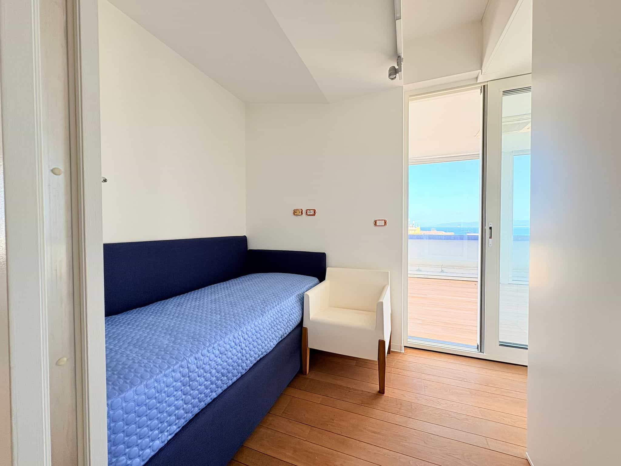 Un letto singolo con testiera blu in una stanza moderna con pareti bianche e pavimento in legno, vicino a una grande finestra che offre vista sul mare e sulla terrazza all'aperto.