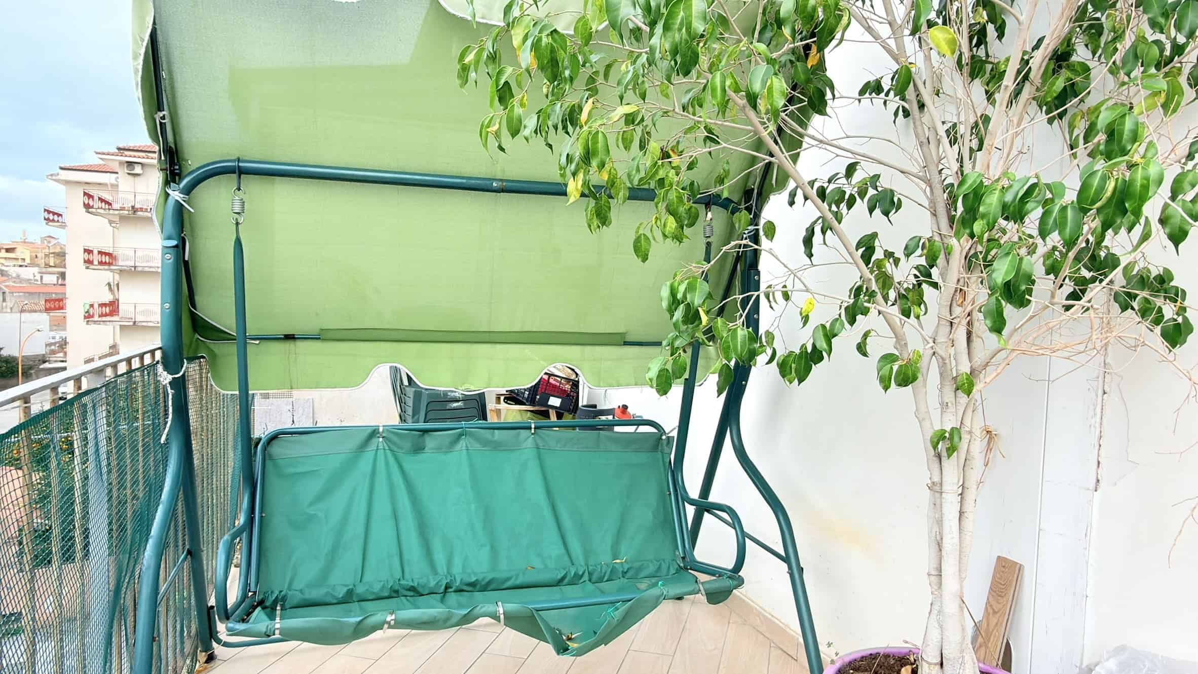 Panchina da giardino con copertura verde, situata su un balcone con vista urbana, circondata da piante verdi e un albero, ideale per relax e momenti di tranquillità all'aperto.