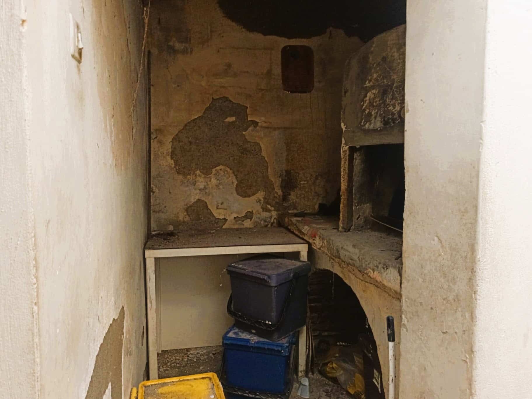 Spazio interno di una vecchia cucina rustica con pareti in pietra e muratura, forno a legna e scaffali in pietra, ambiente da ristrutturare.