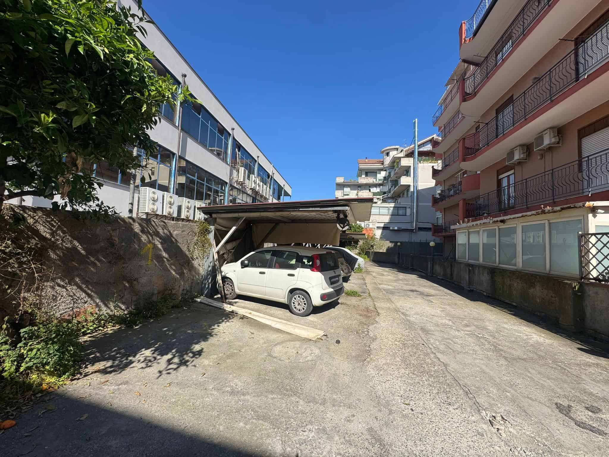 Auto in sosta coperta in un cortile condominiale, tra edifici residenziali moderni, sotto un cielo sereno.