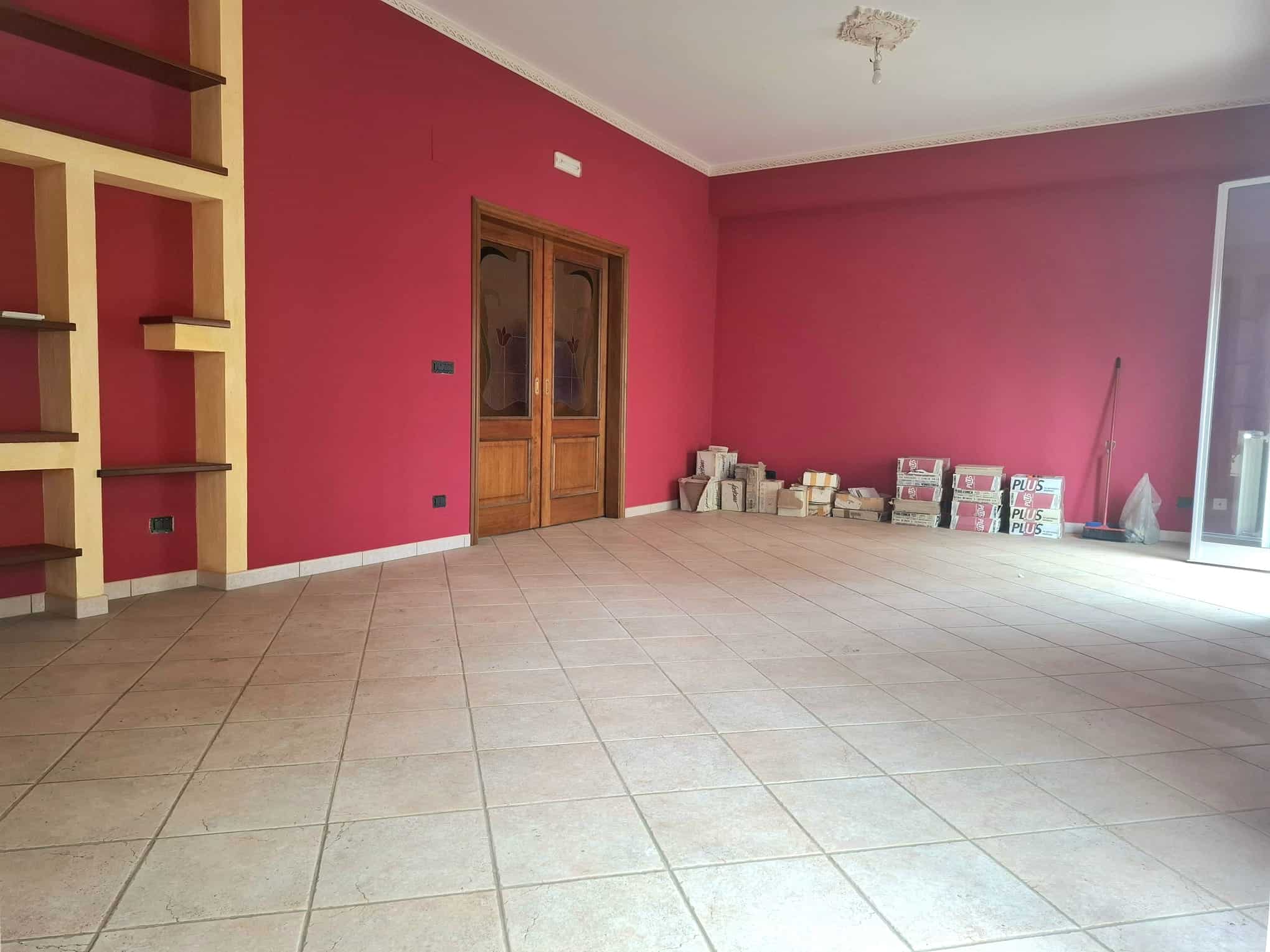 Ampio soggiorno vuoto con pareti rosa e pavimento in piastrelle beige, con scaffali in legno e porta in legno massiccio, illuminato da luce naturale da una grande finestra o porta finestra.