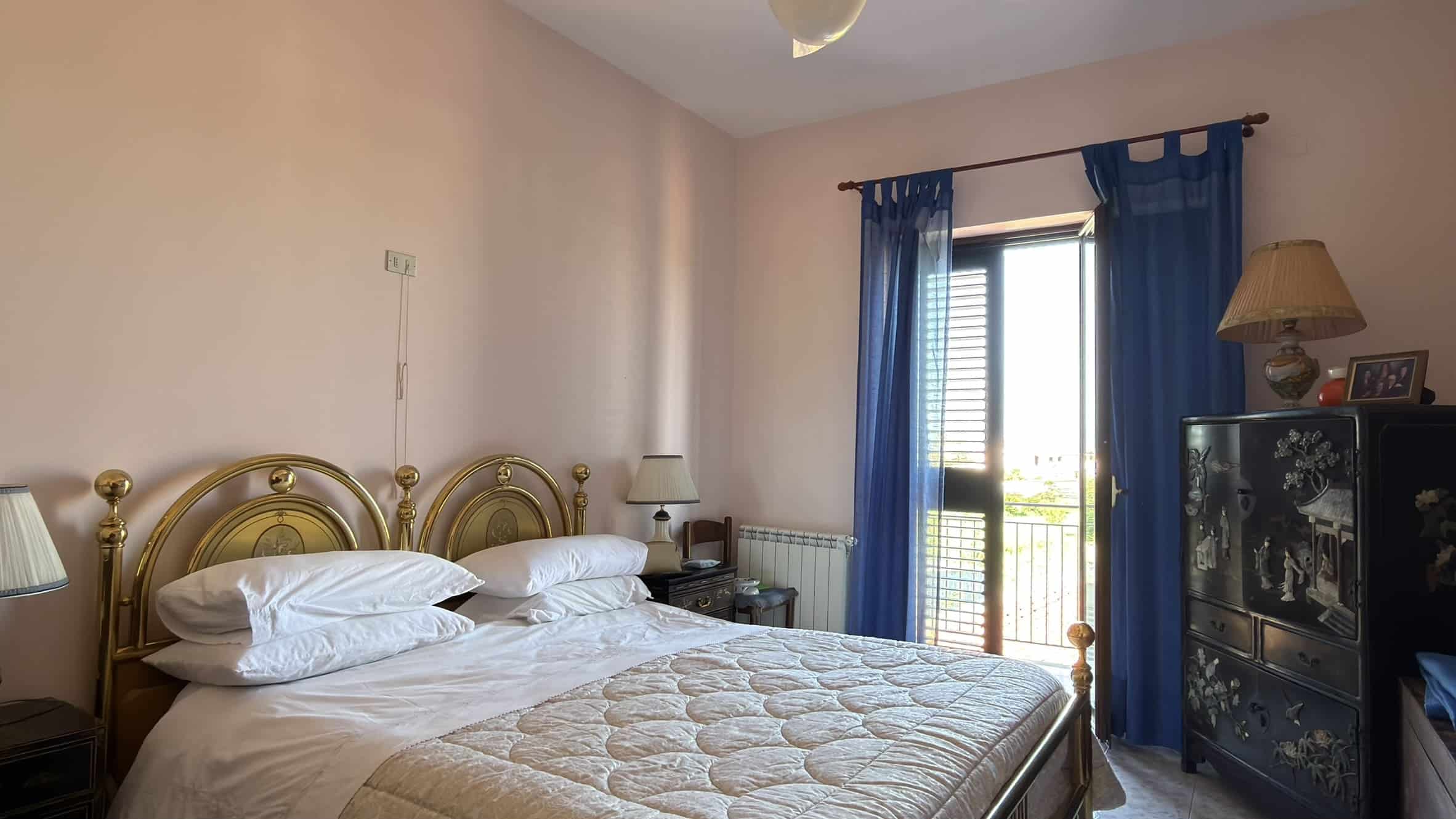 Camera da letto con letto matrimoniale, comodini e accessori, illuminata dalla luce naturale attraverso una porta finestra con tende blu.