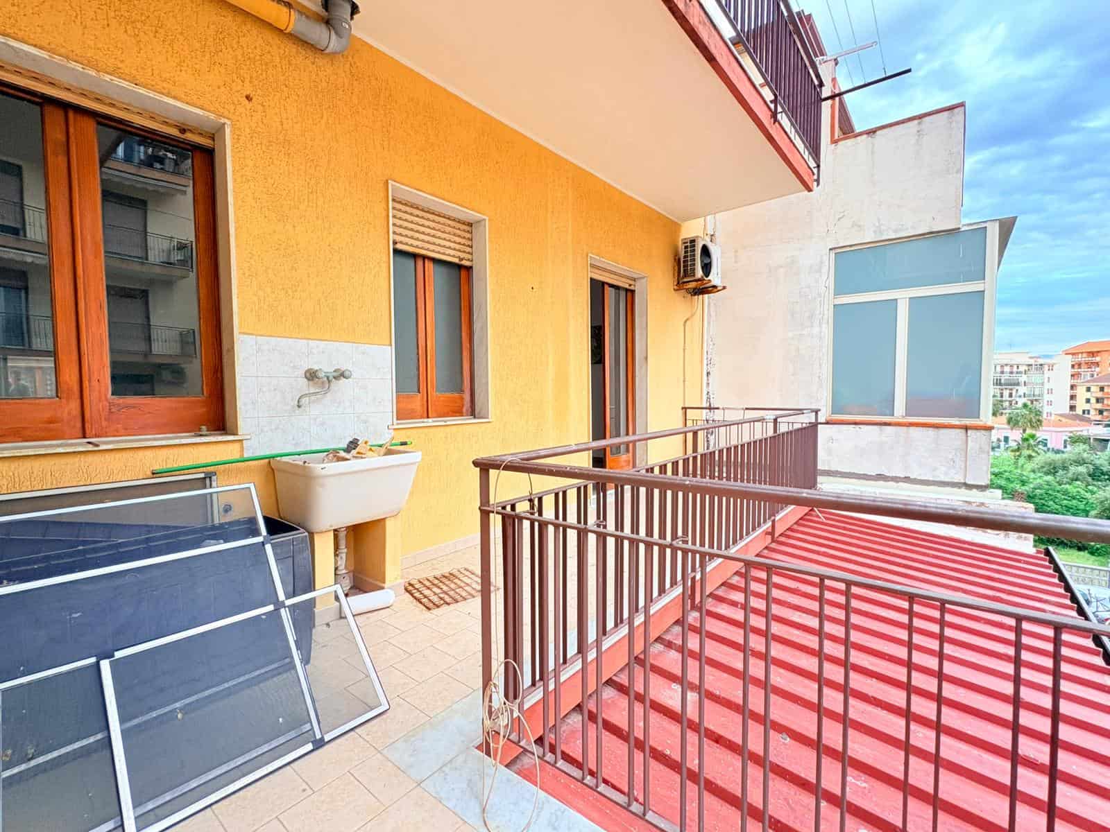Panchetta di un balcone di un appartamento urbano con vista sulla città, con stendibiancheria e lavandino esterno, pareti gialle e finestre con tapparelle, in un ambiente residenziale vivace.