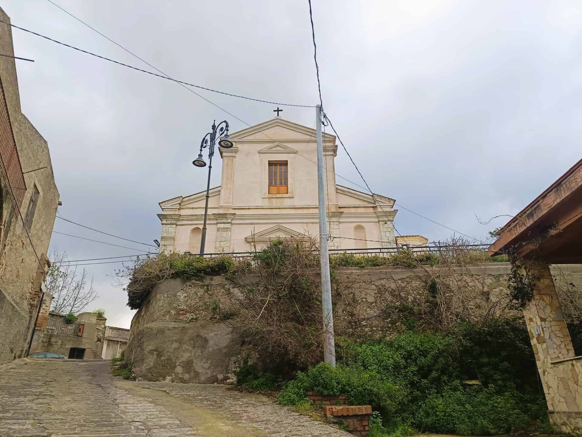 Cattura di una chiesa storica situata su una collina, con un'architettura classica e dettagli decorativi, visibile da una strada in pendenza con lampioni e vegetazione circostante.