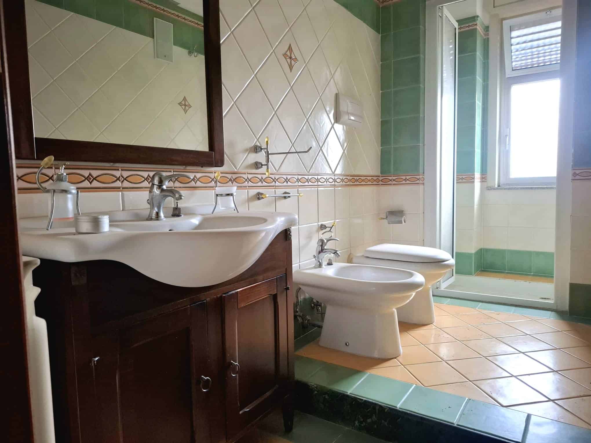 Lavabo e bidet in bagno con piastrelle verdi e beige, finestra luminosa e doccia in background, stile classico e funzionale.