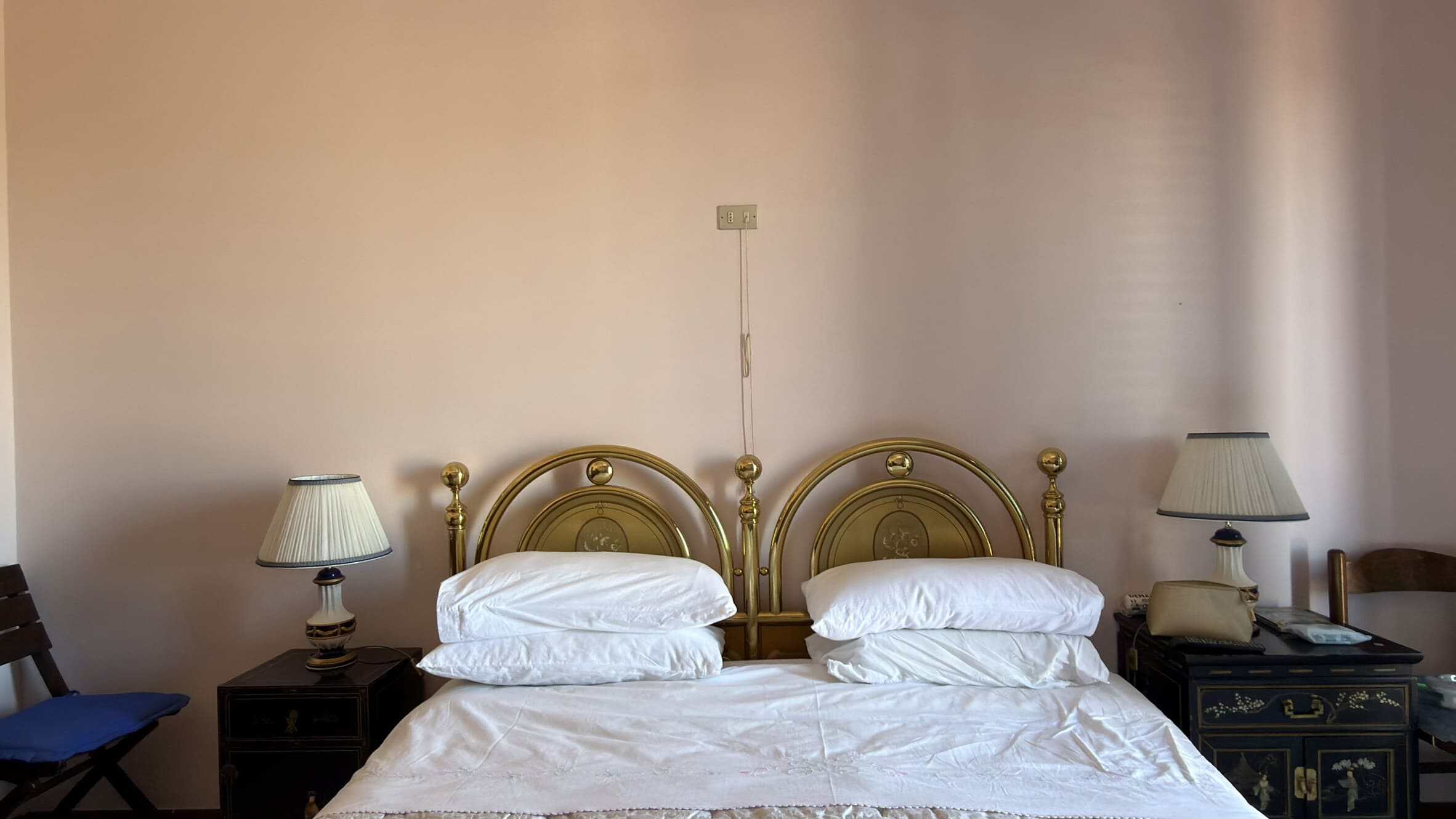 Letto matrimoniale con testiera in metallo dorato, due comodini con lampade da lettura, in una stanza con pareti beige e arredamento classico.