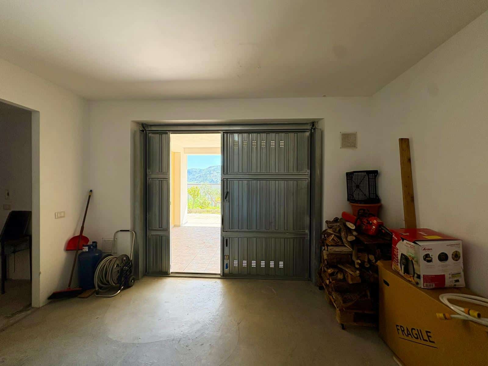 Ampia stanza con porta scorrevole in metallo che si apre su un balcone con vista panoramica sulle montagne, arredamento minimalista, oggetti vari e materiali da lavoro accatastati a destra, pareti bia.