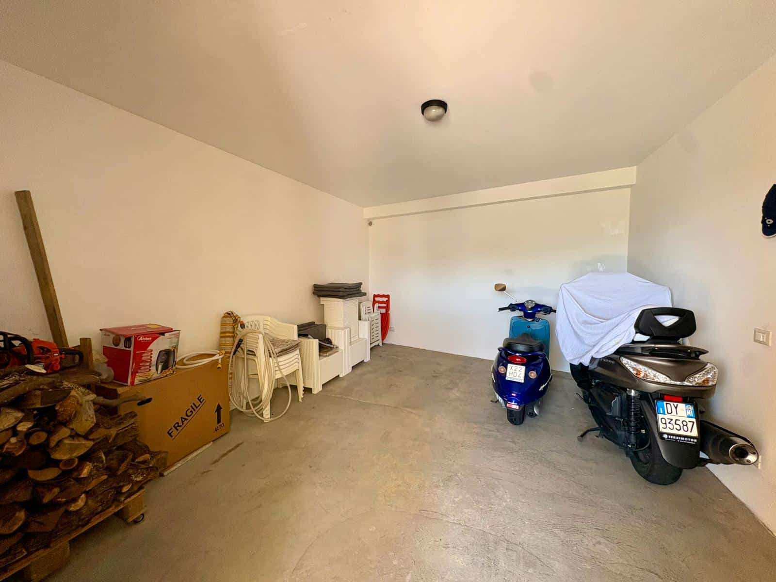 Garage spazioso con scooter, moto e legna da ardere, spazio di stoccaggio e scaffali, ambiente luminoso e ben organizzato.