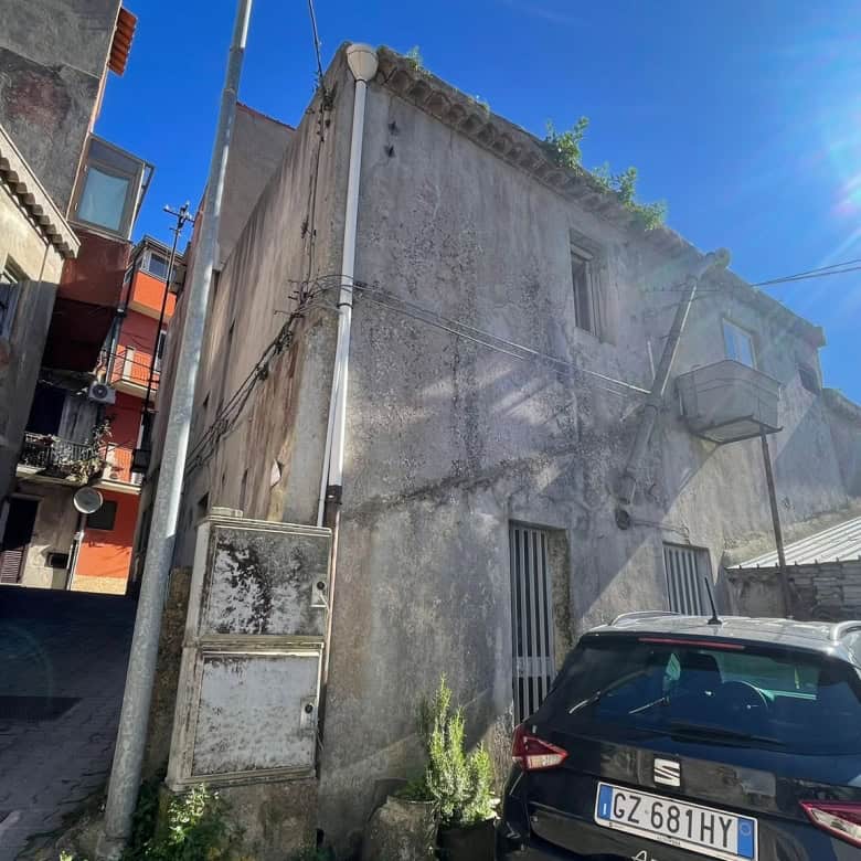 Vecchio edificio in pietra con pareti grigie e finestre piccole, situato in un vicolo urbano soleggiato con auto parcheggiata davanti, sotto un cielo blu con sole brillante.