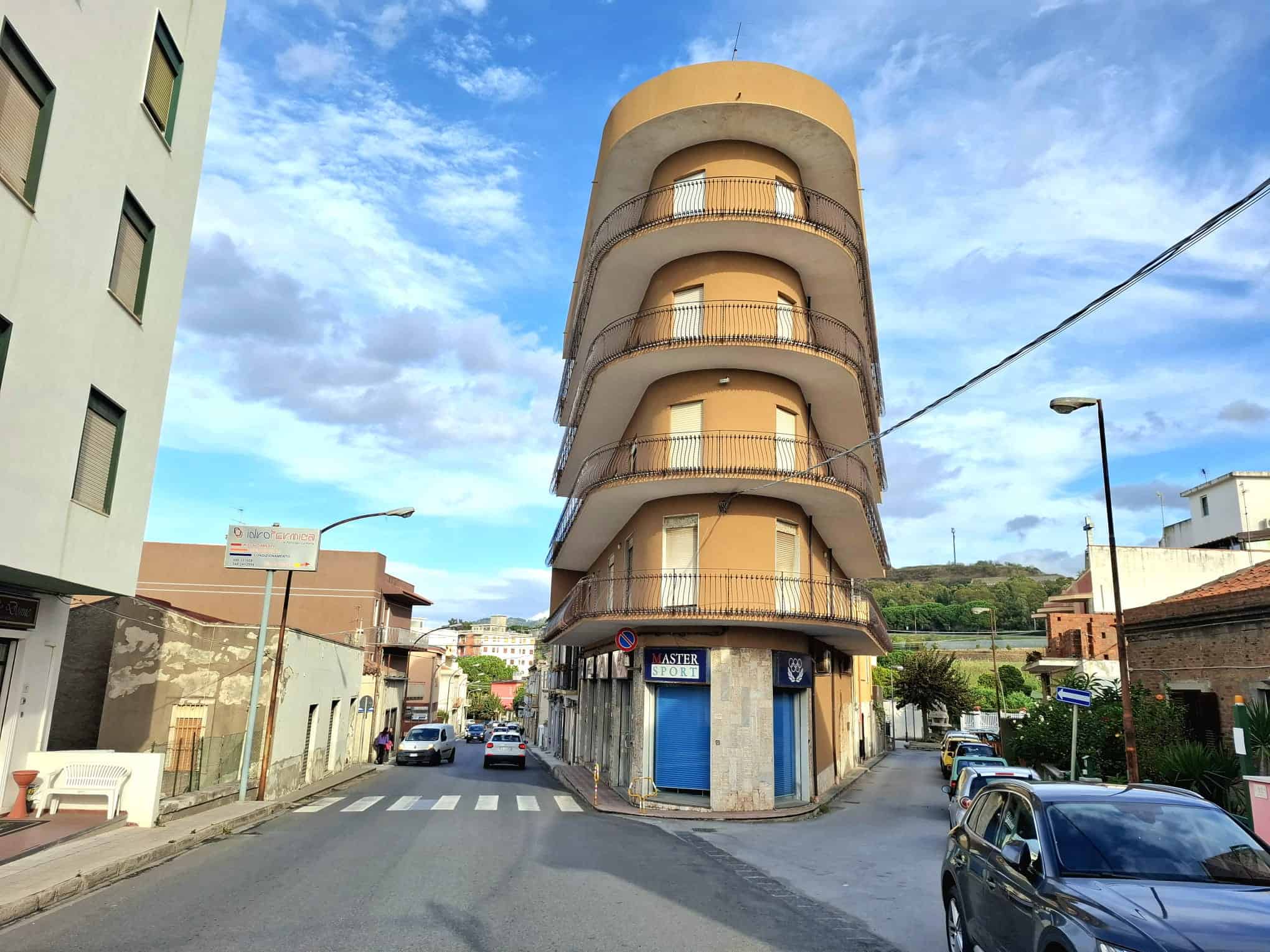 Edificio residenziale a quattro piani con balconi rotondi, situato in un'area urbana con strada trafficata e veicoli parcheggiati, sotto un cielo nuvoloso.