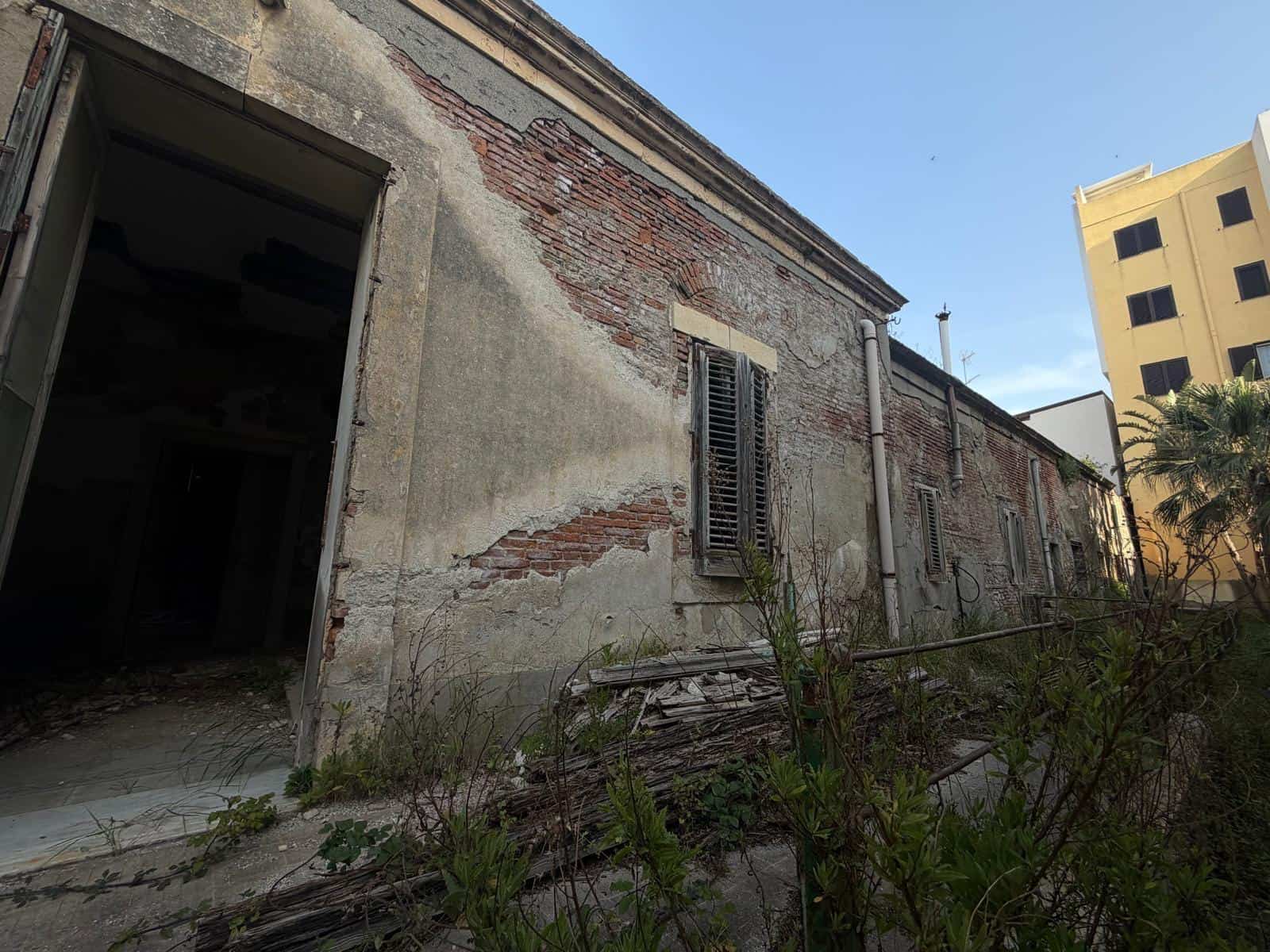 Abbandono di edificio storico in rovina con pareti in pietra e mattoni, finestre con persiane in legno, vegetazione selvaggia e cielo azzurro, rappresentando un esempio di architettura decadente e deg.