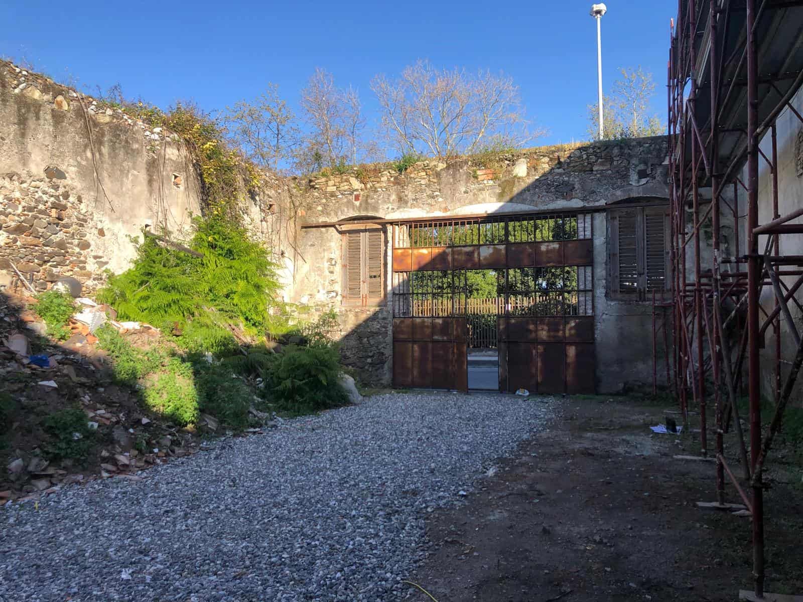 Cancello in ferro battuto con pareti in pietra e accesso a un cortile interno, in un'area esterna con cielo sereno e vegetazione circostante.