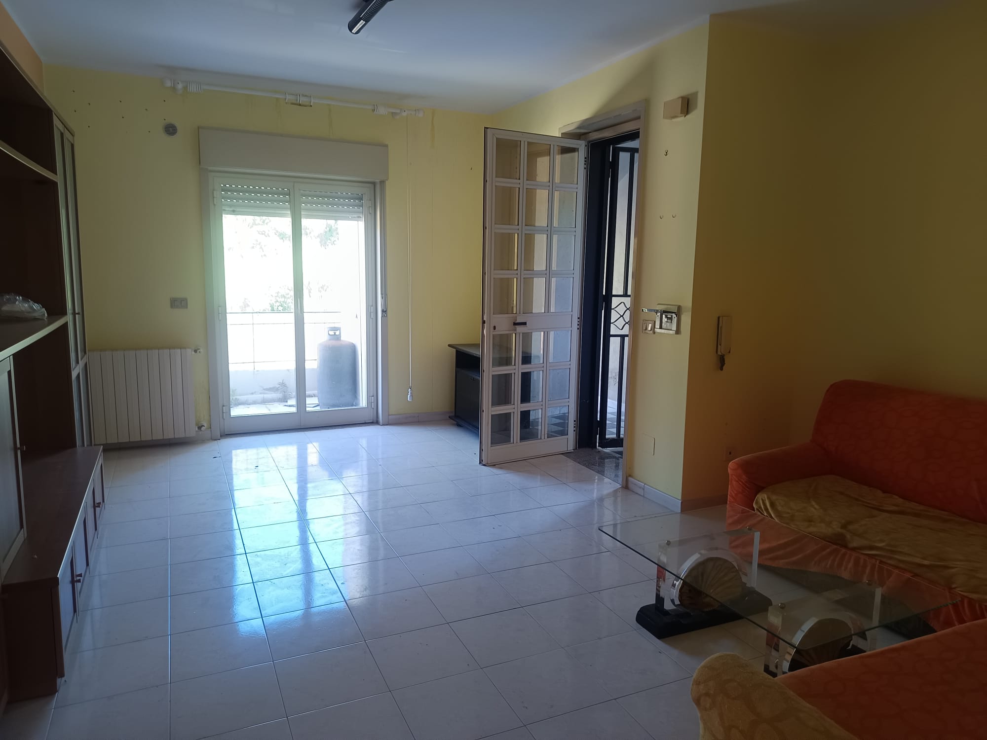 Interno di un soggiorno luminoso con porta finestra che dà su un balcone, pareti gialle, divano arancione e tavolino in vetro, ideale per affitti o vendita immobili.